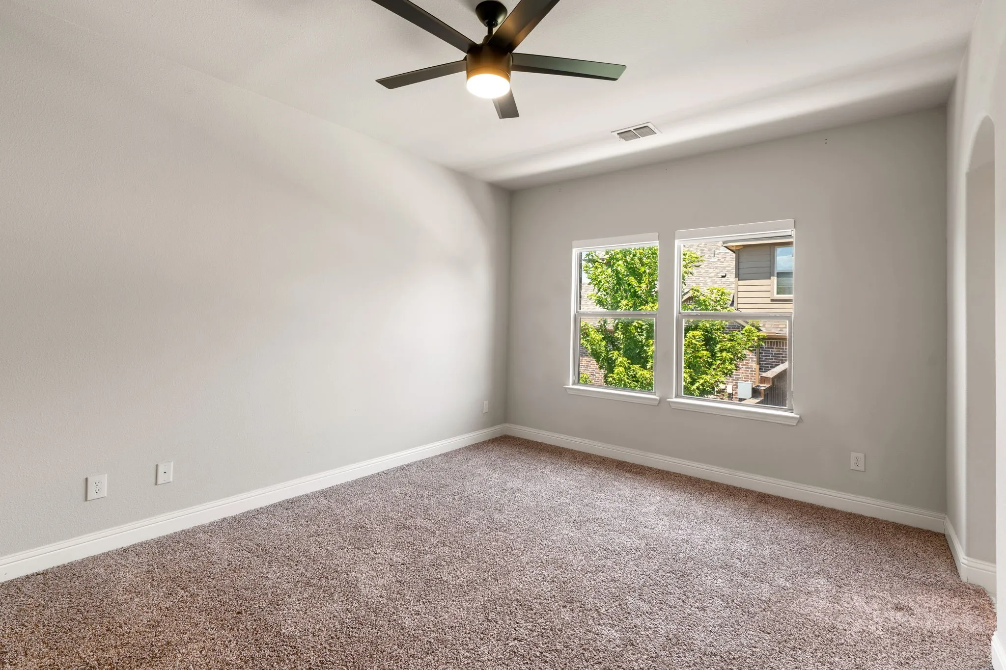 Neutral wall paint, carpet flooring, two bright windows, ceiling fan with integrated lighting, and an arched entryway