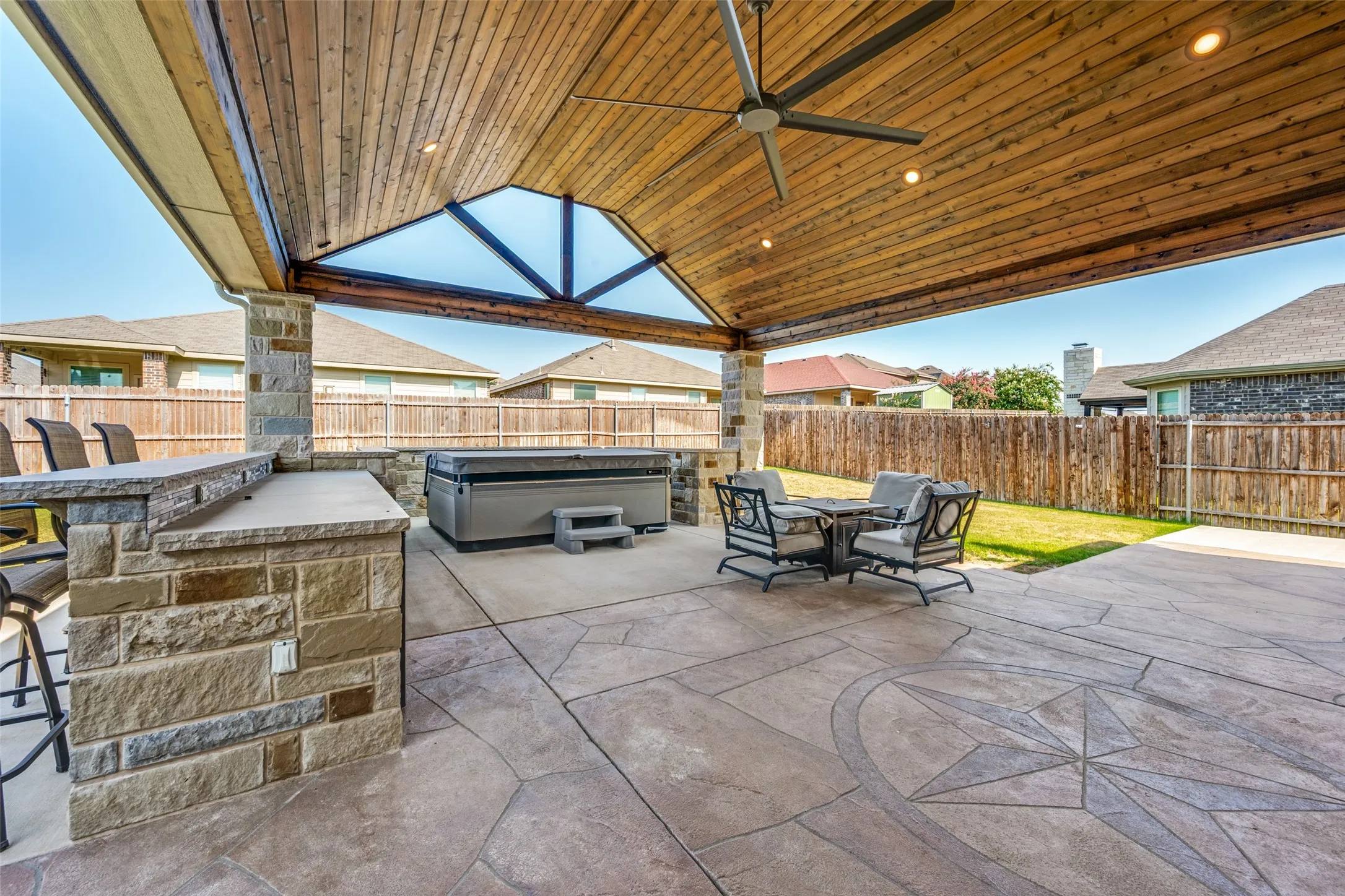 Expansive covered patio featuring a wood-finish ceiling with integrated lighting, stone fireplace with built-in seating ledge, and stamped concrete flooring