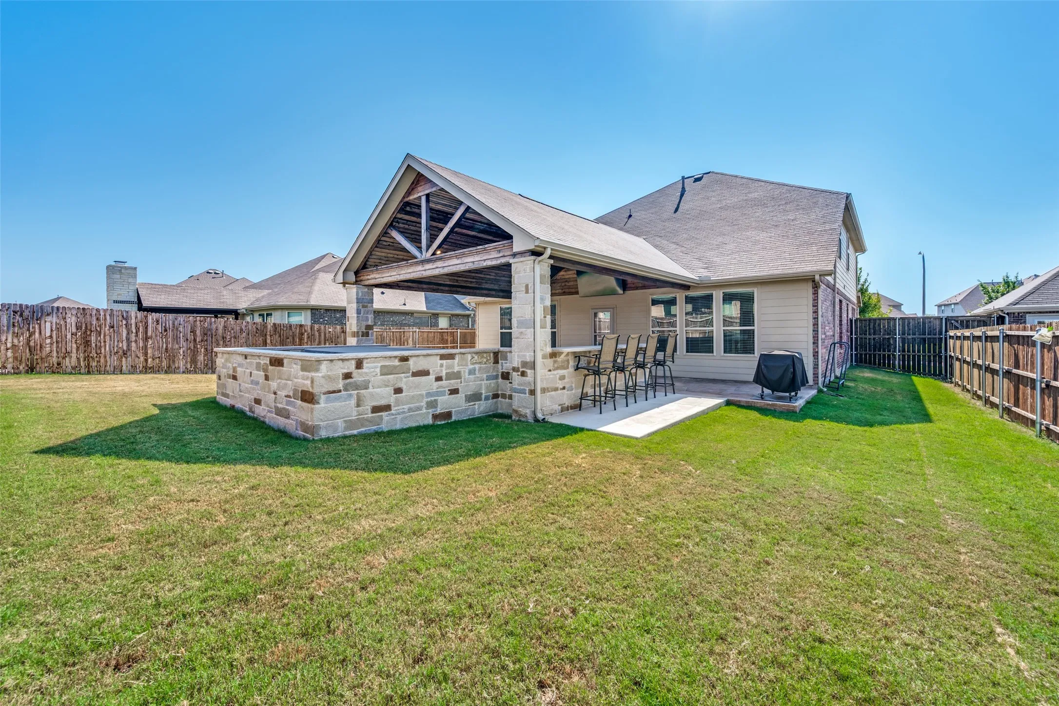 Expansive backyard oasis featuring a covered patio with stone-veneer accents, pitched roof with exposed beam detailing, and mature landscaping with manicured lawn and wooden privacy fencing