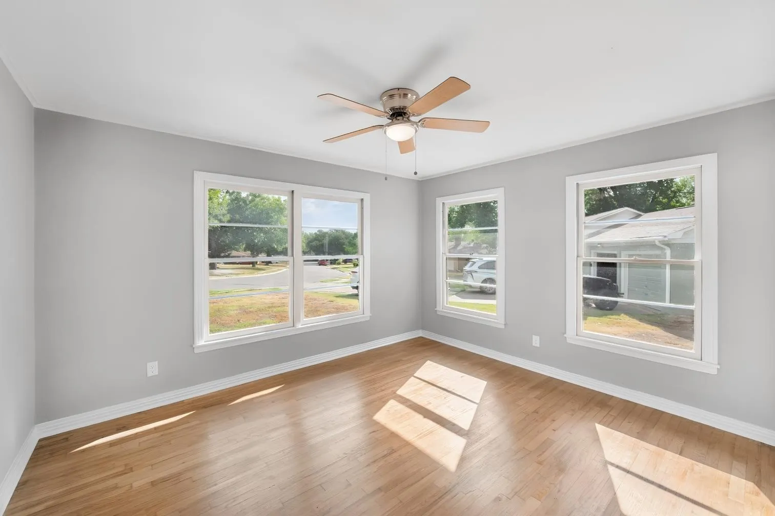 Light gray walls, wood-finish flooring, two large windows, a single window, and a ceiling fan with integrated lighting