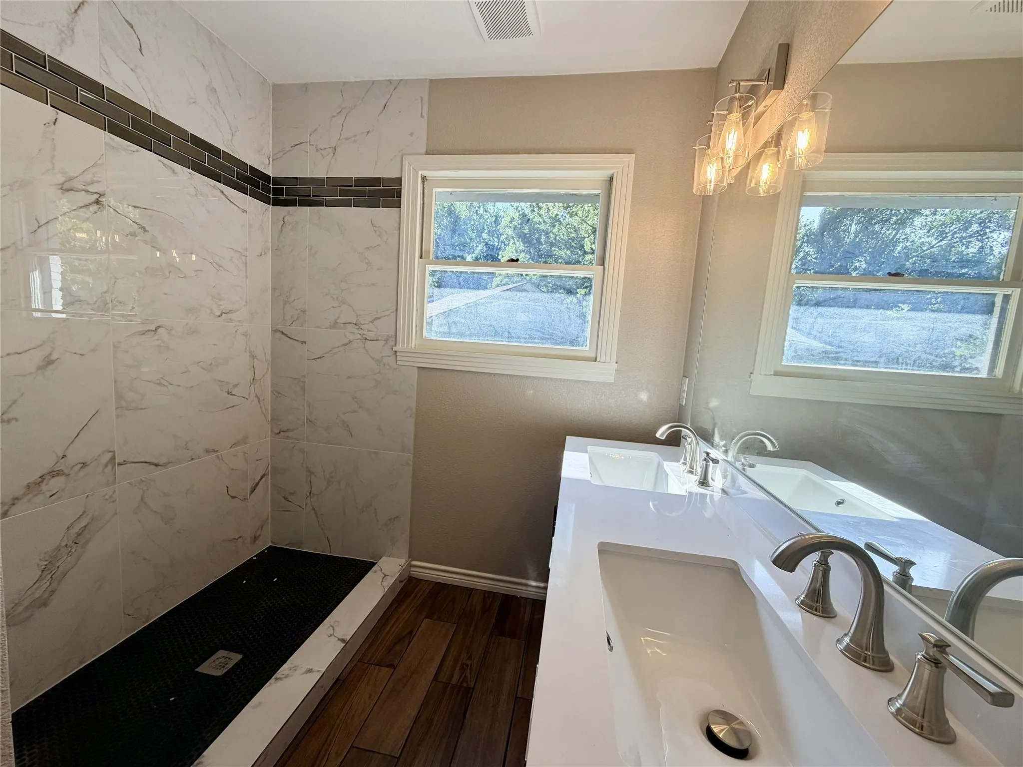 Spa-inspired bathroom featuring a walk-in shower with marble-look tile surround and dark accent stripe, wood-finish ceramic tile flooring, dual vanity with white quartz countertop and chrome fixtures, recessed lighting, and natural light
