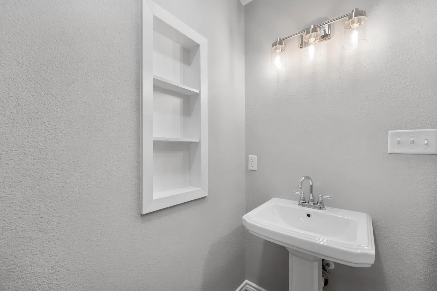 Pedestal sink with chrome-finish faucet, built-in wall shelving, and a three-light vanity fixture