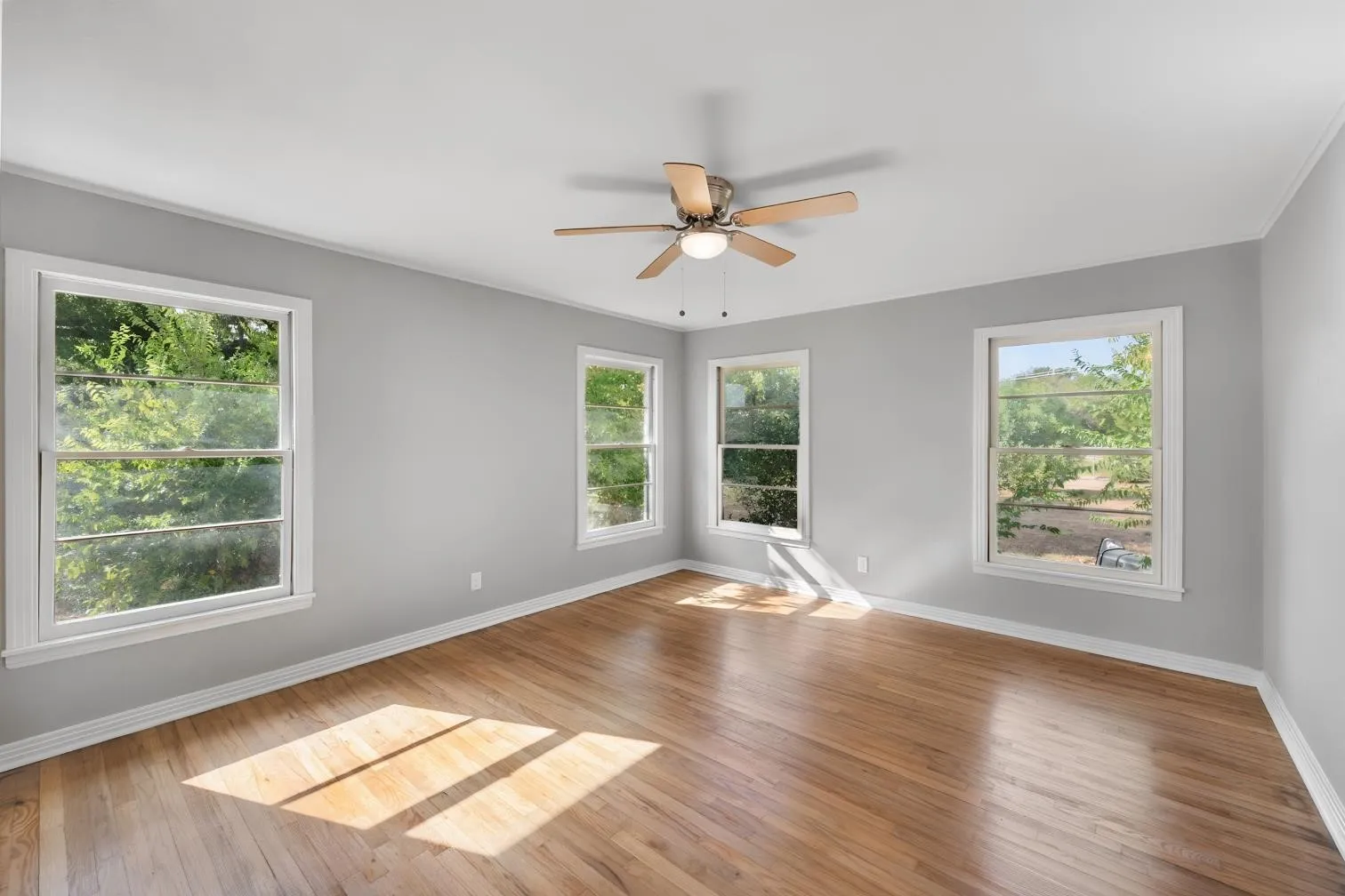 Hardwood flooring, light gray wall paint, white trim, three windows, and a ceiling fan with integrated lighting