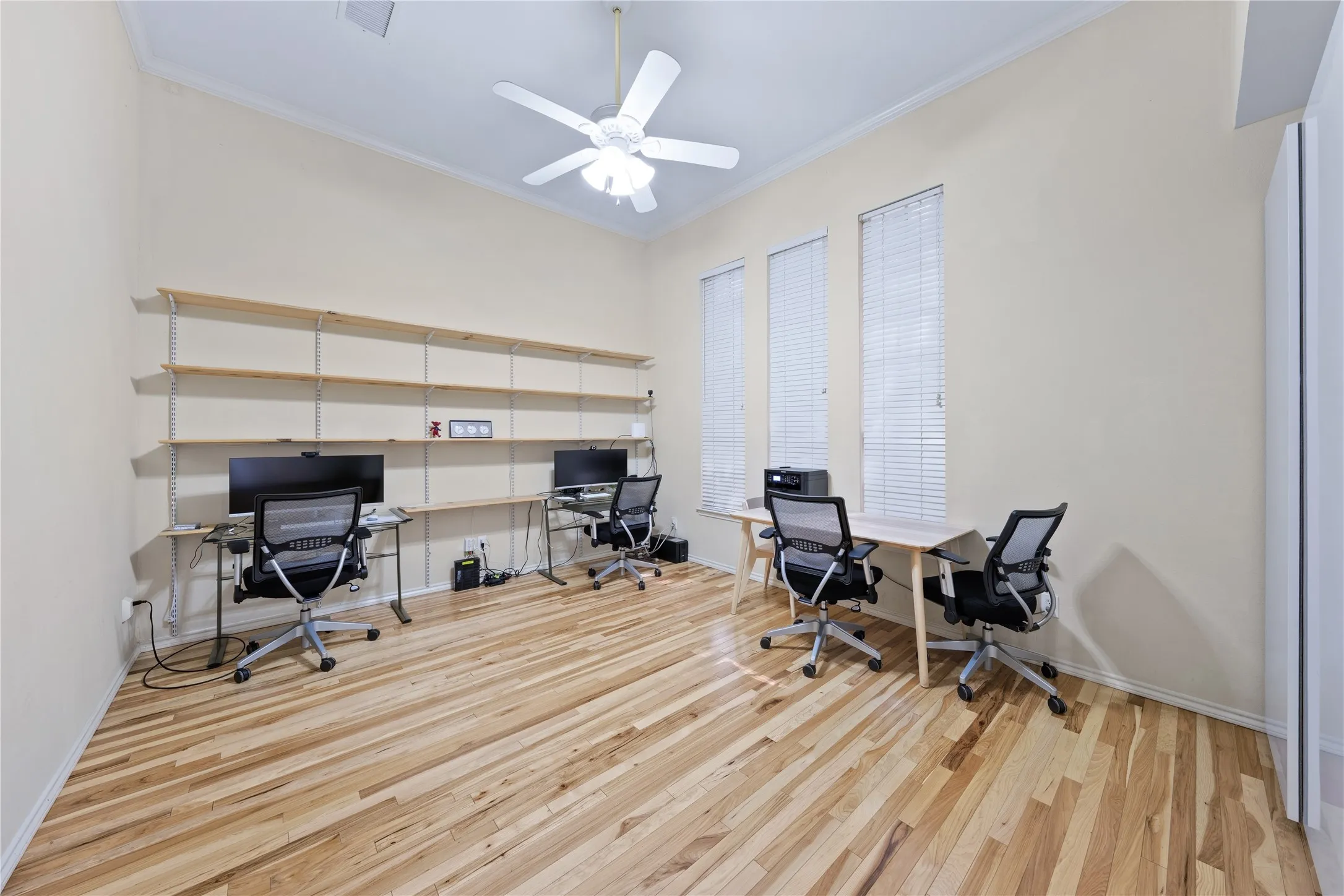 Room featuring natural wood flooring, three vertical windows with blinds, a ceiling fan, and built-in wall shelving