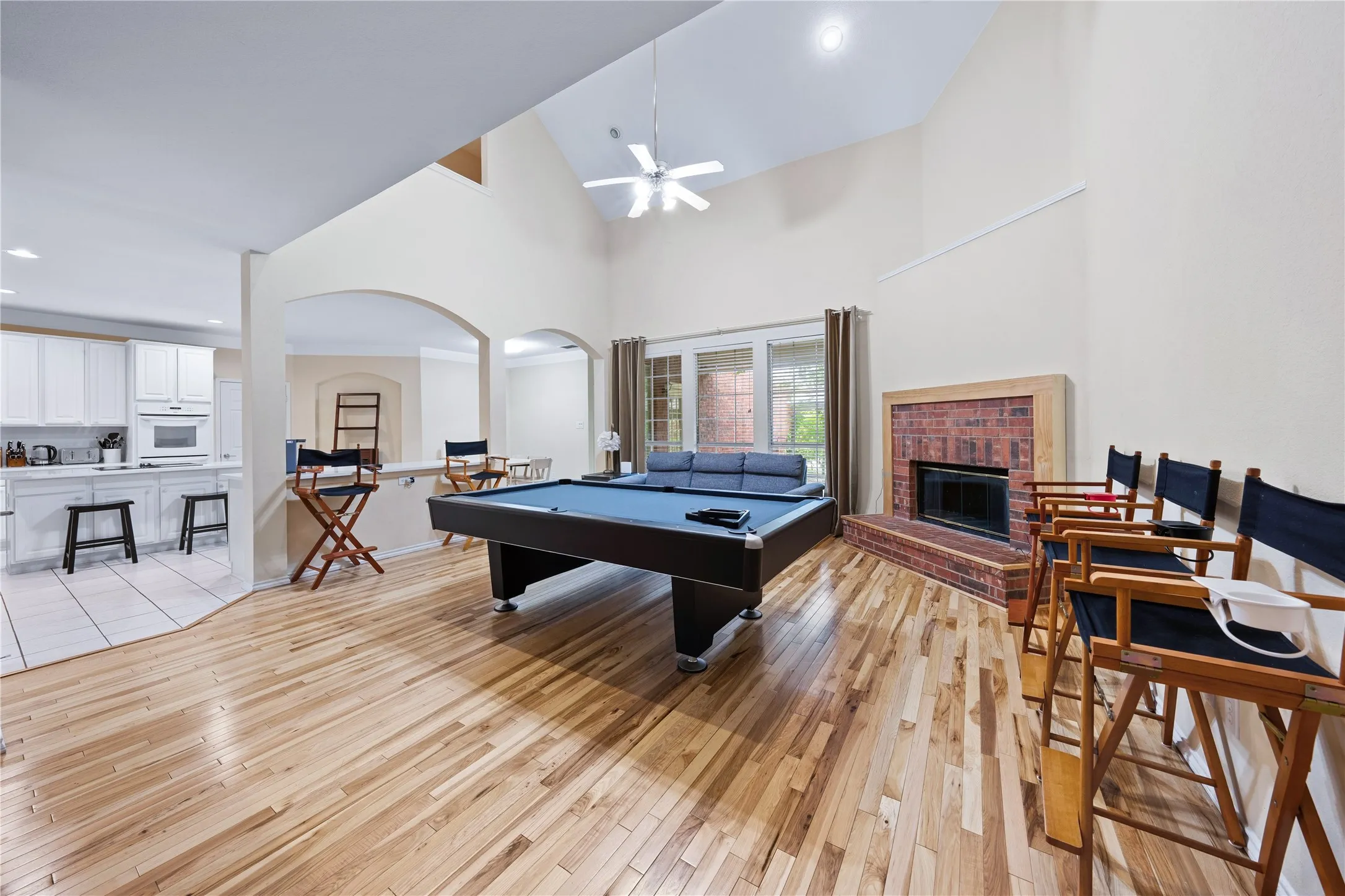Vaulted ceiling with ceiling fan, hardwood flooring, brick-surround fireplace, arched doorways, and a kitchen with white cabinetry and tile flooring