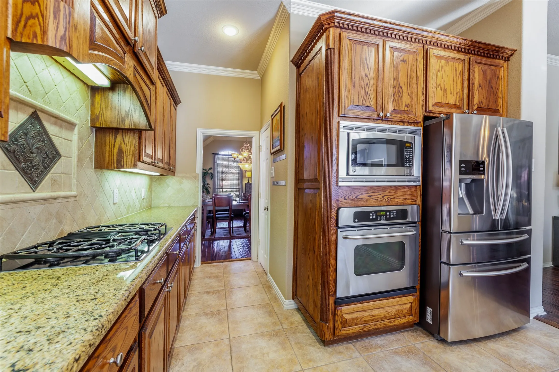 Kitchen with stainless steel appliances, tasteful backsplash, brown cabinetry, ornamental molding, and light stone countertops