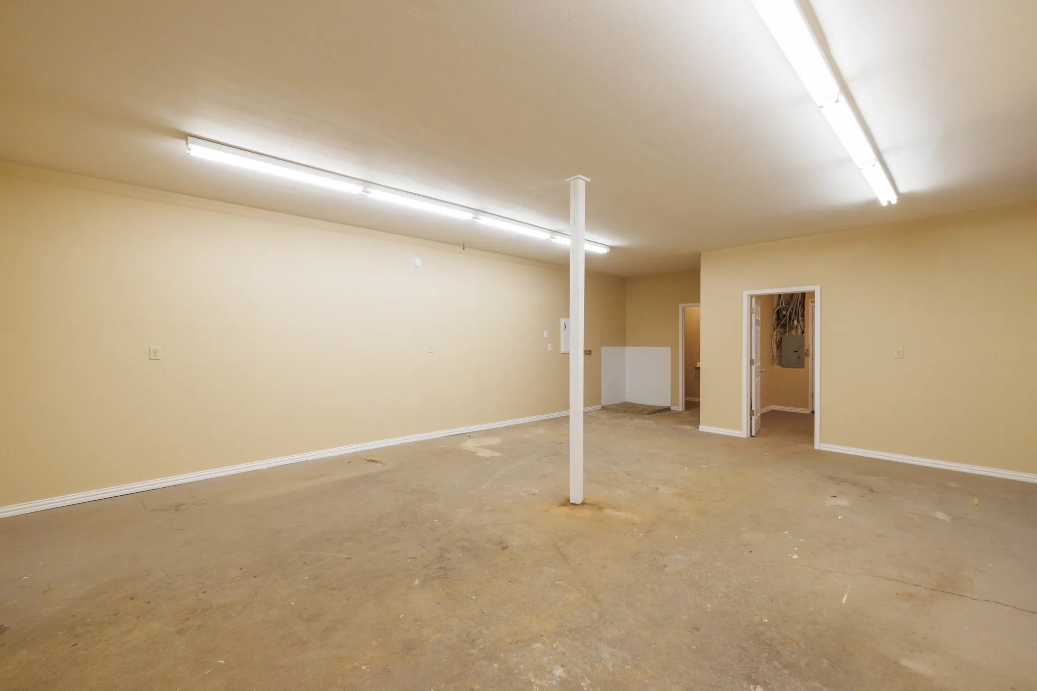 Spacious room featuring concrete flooring, neutral wall color, overhead fluorescent lighting, and multiple doorways