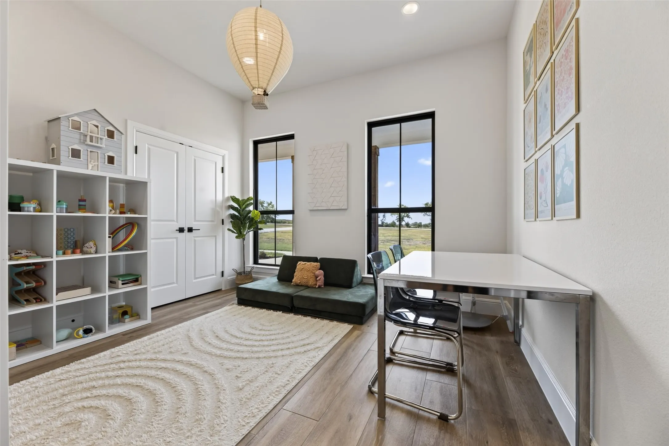 Bright, light-filled home office featuring wood-finish flooring, white built-in cabinetry with open shelving, a desk with metal frame and glass top, black upholstered seating, large windows with black frames, a woven pendant light fixture, and a geometric patterned area rug