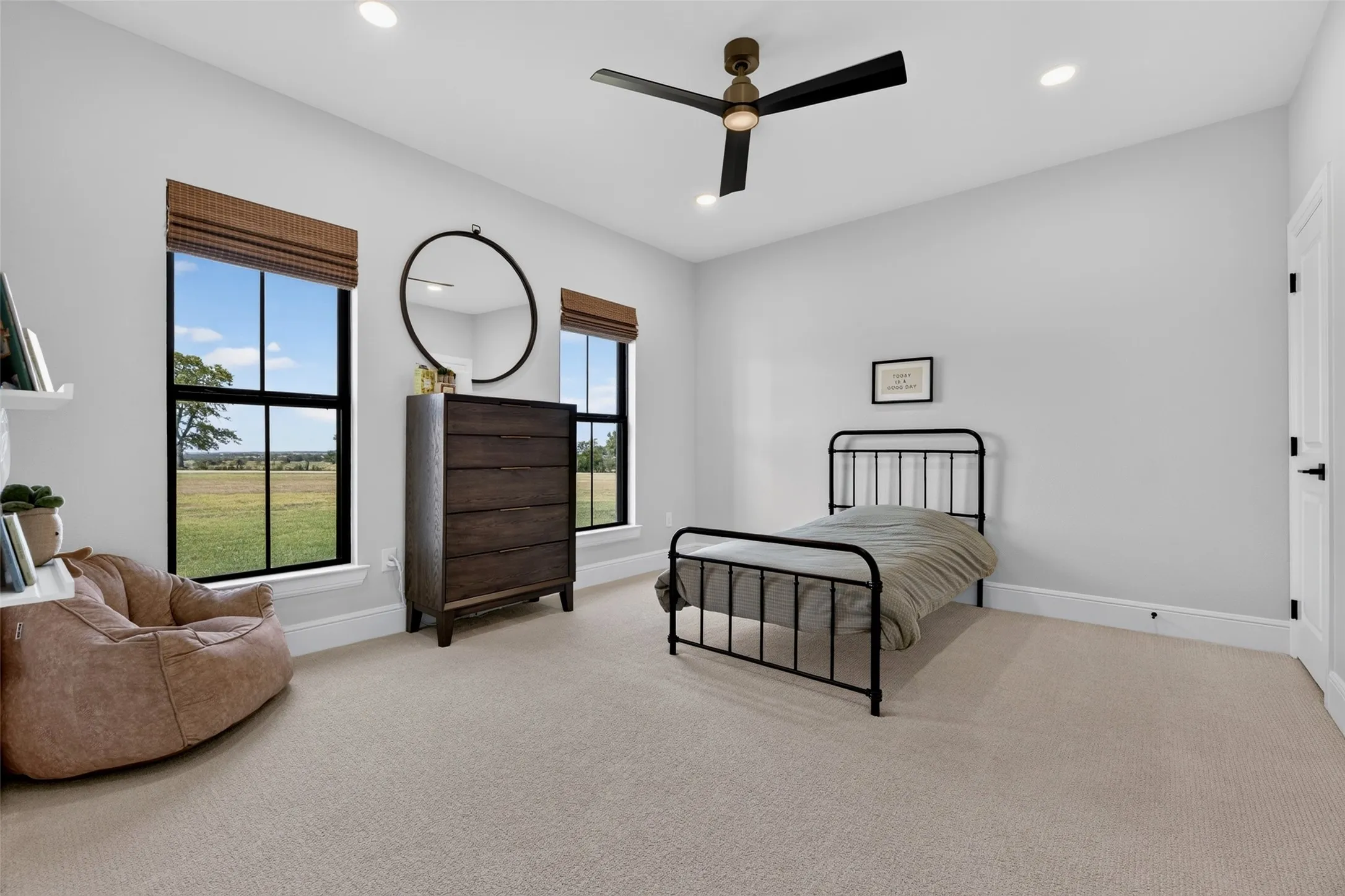 Bright secondary bedroom with dual large windows framing pastoral landscape vistas, neutral carpet flooring, metal bed frame, wood-finish dresser with round mirror, ceiling fan with integrated lighting, and recessed ceiling lights