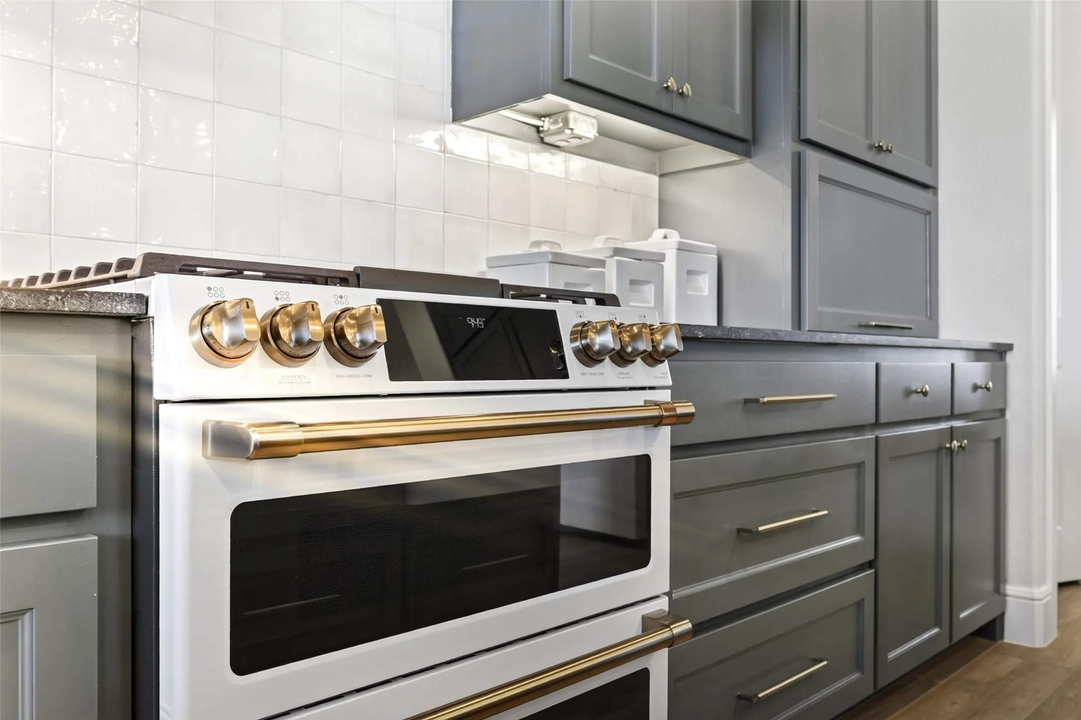 Contemporary kitchen featuring a white and stainless steel dual wall oven with gold hardware accents, five-burner gas cooktop with matching gold trim, gray shaker-style cabinetry with brushed metal handles, white subway tile backsplash, and wood-finish flooring