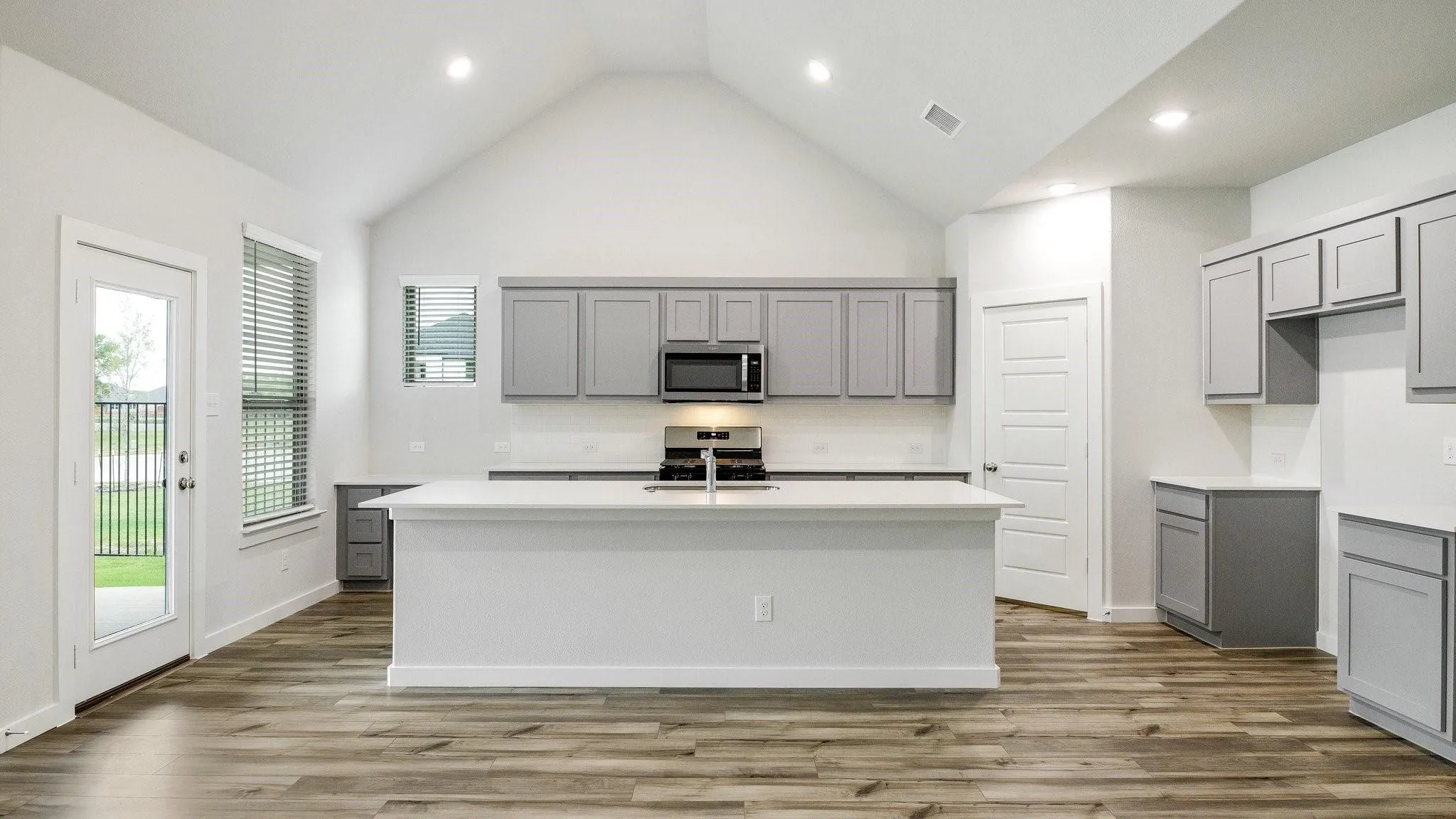 Open-concept kitchen featuring a large island with white cabinetry, stainless steel appliances, wood-finish flooring throughout, vaulted ceiling with recessed lighting, and direct access to outdoor space via French doors