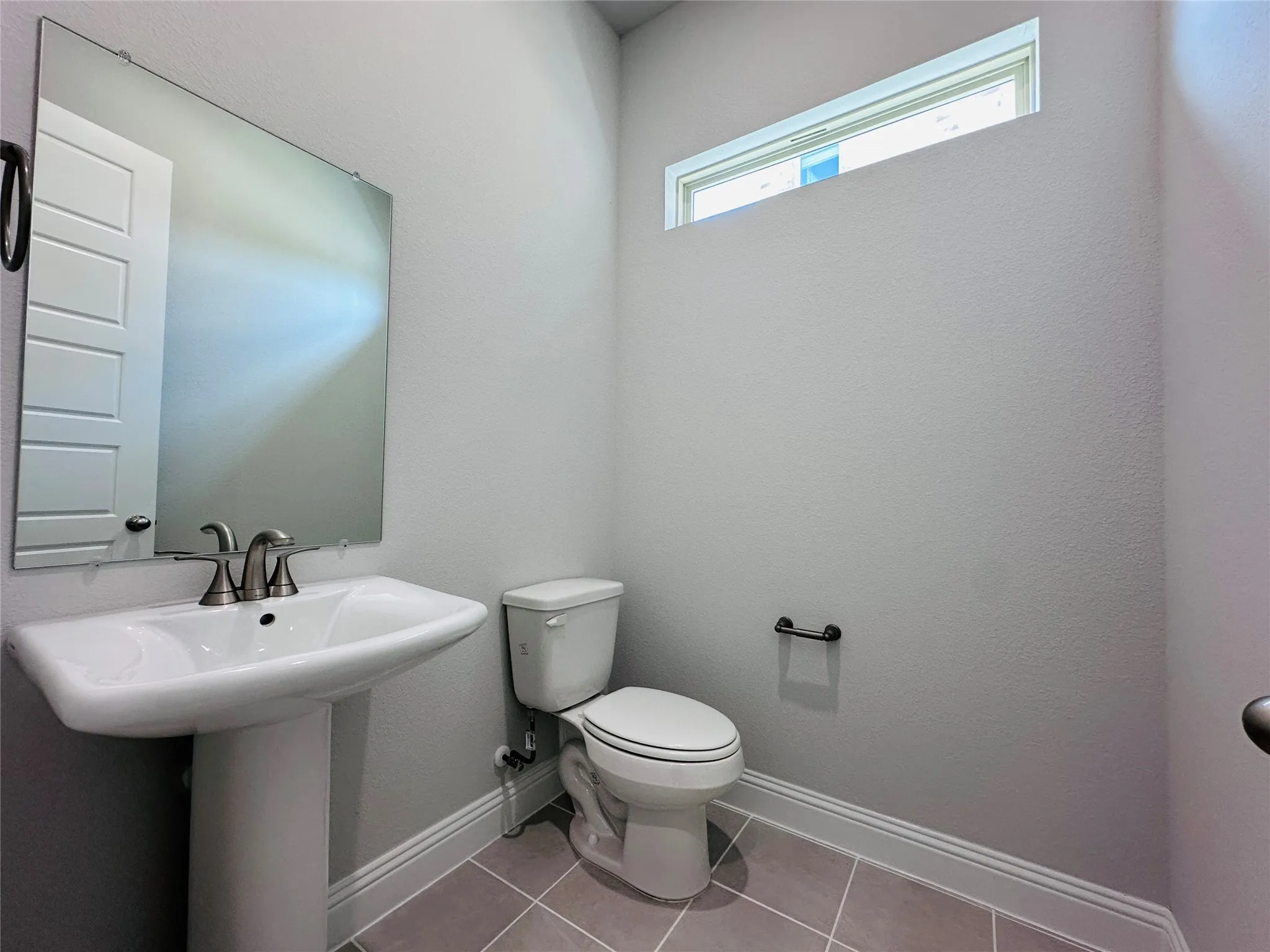 Powder room featuring a pedestal sink with chrome faucet, ceramic toilet, tile flooring, and a transom window providing natural light