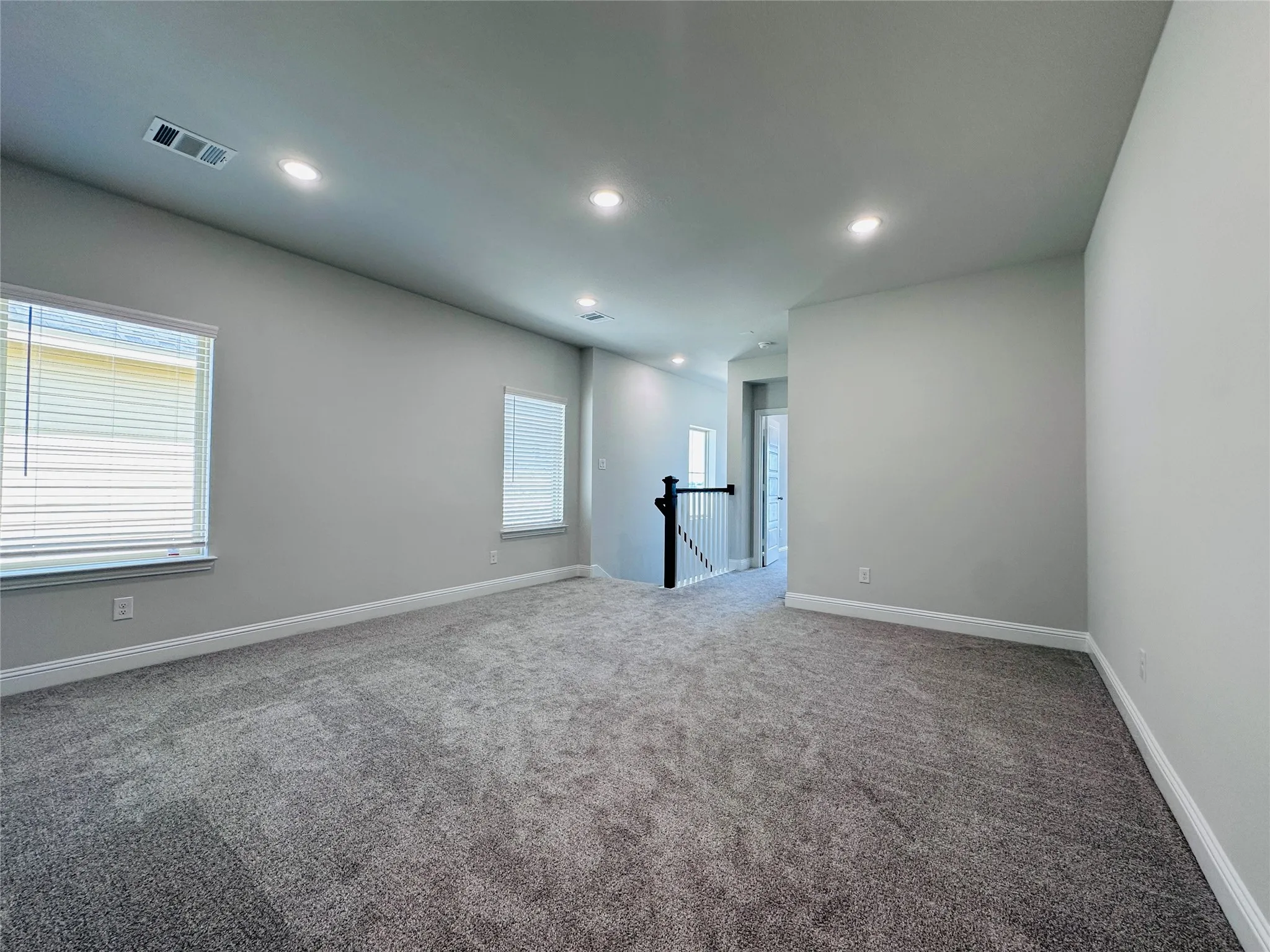 Spacious room featuring neutral carpeting, multiple windows with blinds, recessed ceiling lighting, and a partial black and white staircase railing