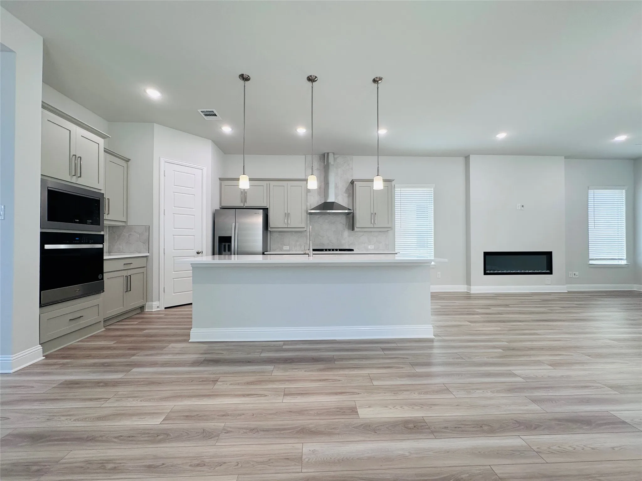 Open-concept kitchen and living area featuring white cabinetry, stainless steel appliances, a large center island with pendant lighting, wood-finish flooring throughout, and recessed ceiling lights