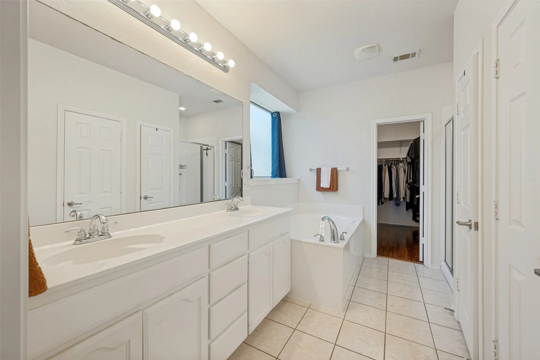 Spa-inspired master bathroom featuring dual vanities with ample storage, a large frameless mirror with integrated lighting, separate soaking tub, tile flooring, and direct access to a walk-in closet