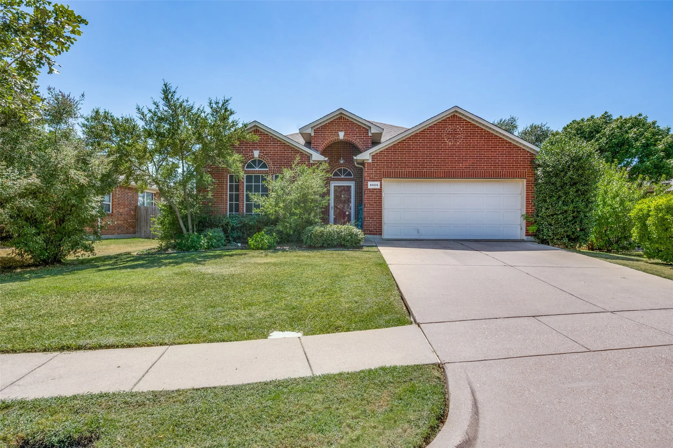 Brick Colonial home with a two-car attached garage, concrete driveway, and manicured front lawn bordered by mature landscaping and established trees throughout the property