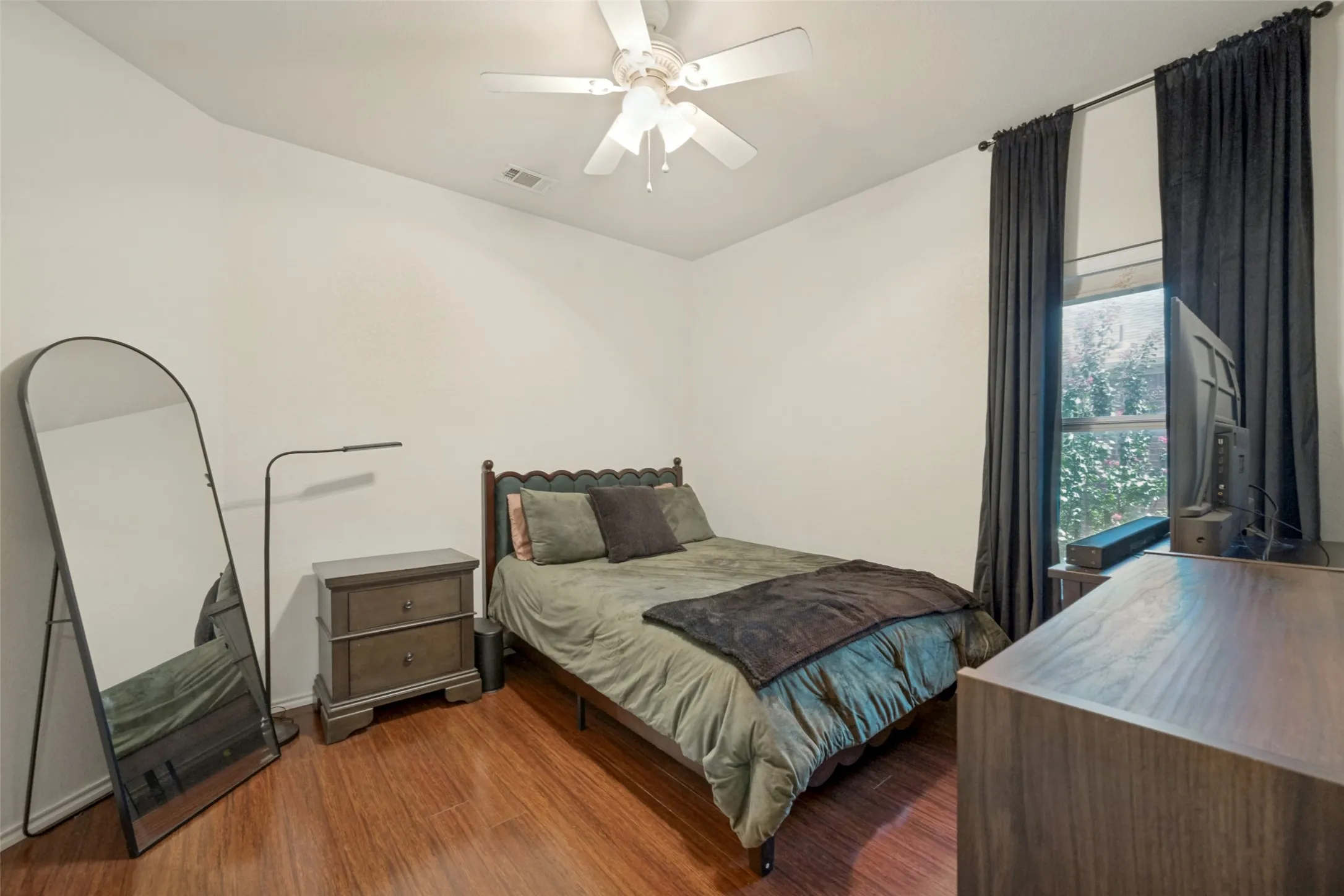 Wood-finish flooring, ceiling fan, white walls, and a window with dark curtains