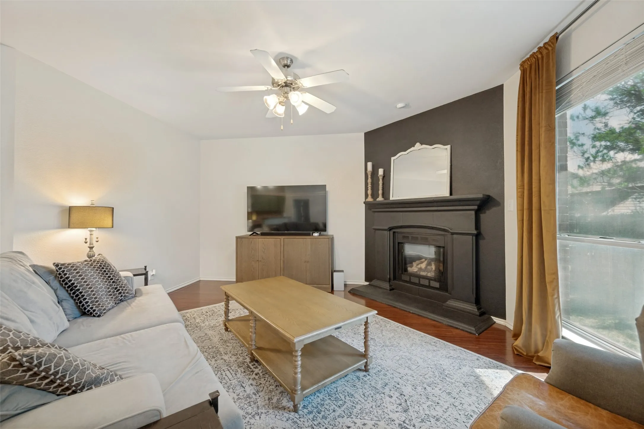 Hardwood-style flooring, gas fireplace with detailed mantel, ceiling fan, and large window with vertical blinds