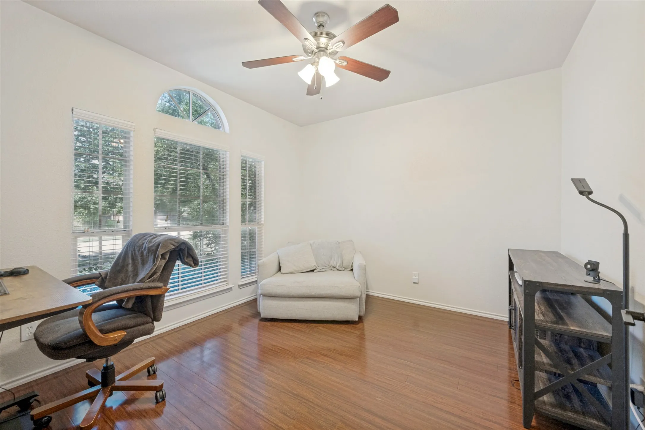 Bright, naturally lit home office featuring expansive windows with plantation shutters, hardwood flooring, ceiling fan with integrated lighting, and ample space for workspace and furnishings