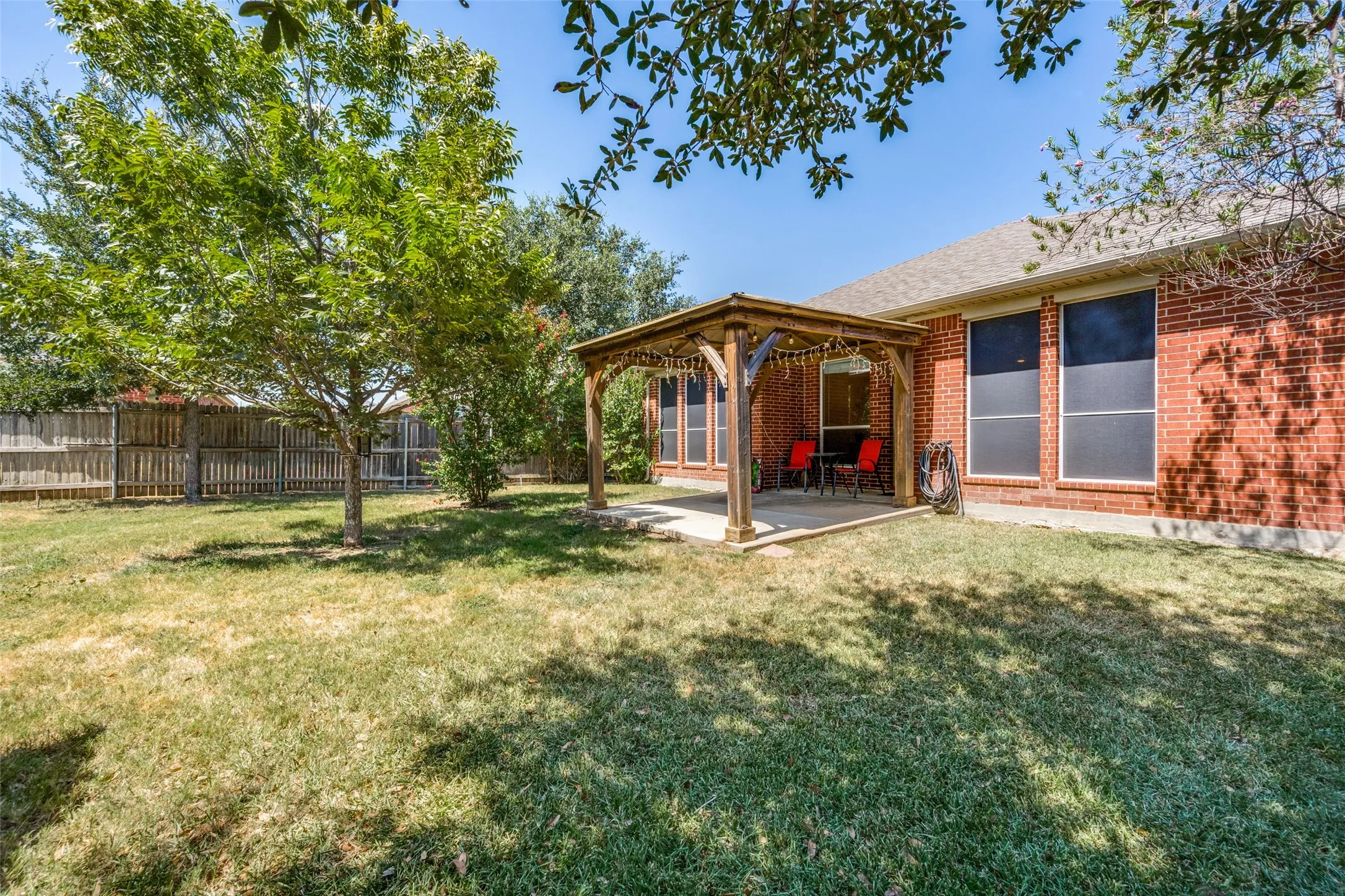 Brick exterior with expansive backyard featuring mature shade trees, manicured lawn, and covered wooden patio structure