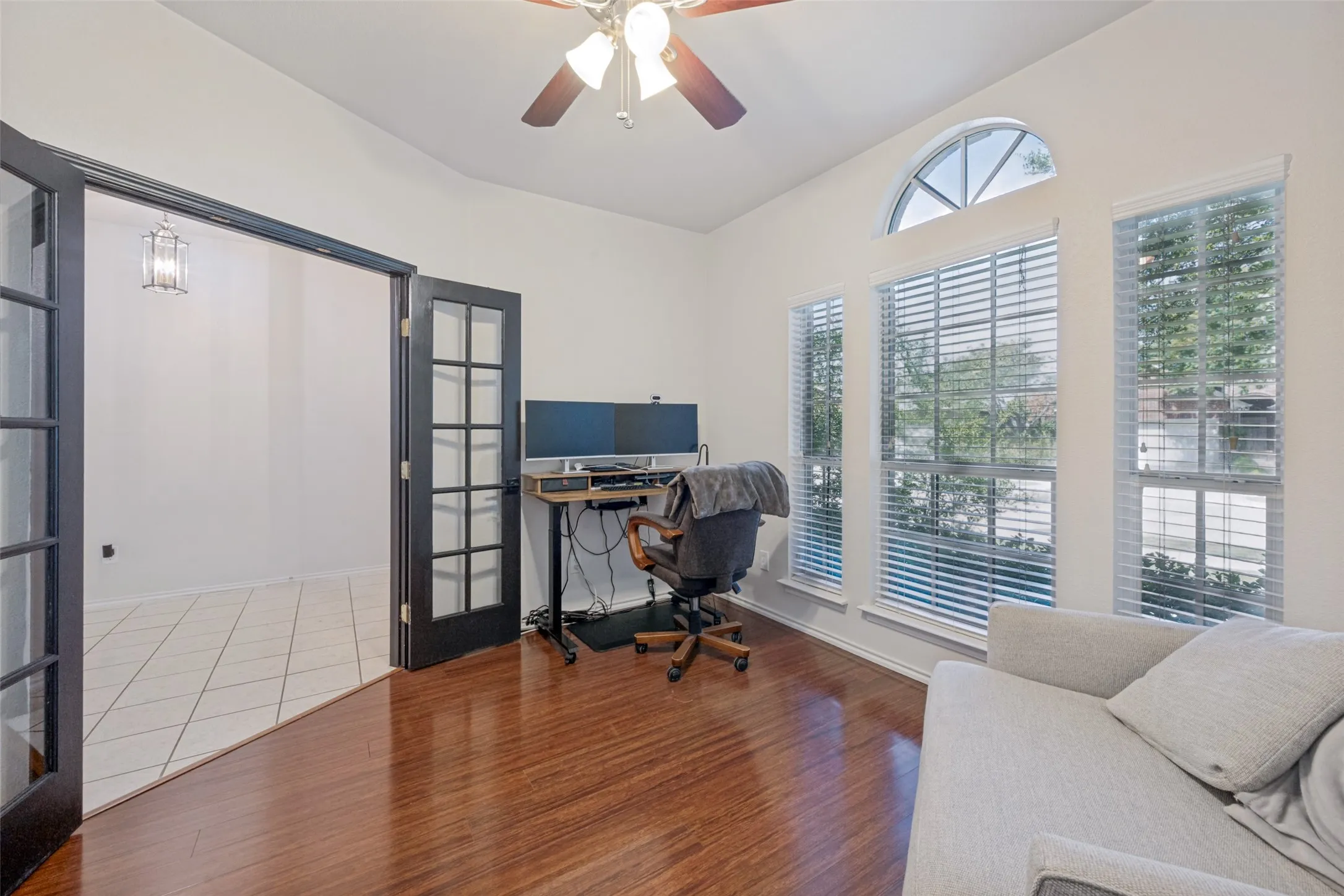 Bright home office featuring hardwood flooring, abundant natural light from multiple windows with plantation shutters, and a ceiling fan with integrated lighting