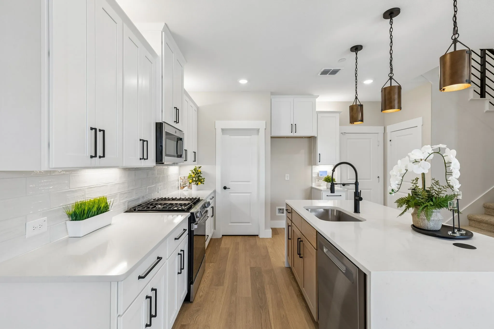 White cabinetry with matte black hardware, stainless steel appliances, light wood-finish flooring, white countertop surfaces, and pendant lighting fixtures
