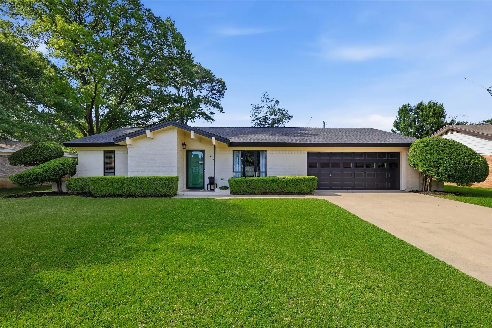 White brick exterior, dark asphalt shingle roof, attached garage with dark panel door, concrete driveway, and established front landscaping