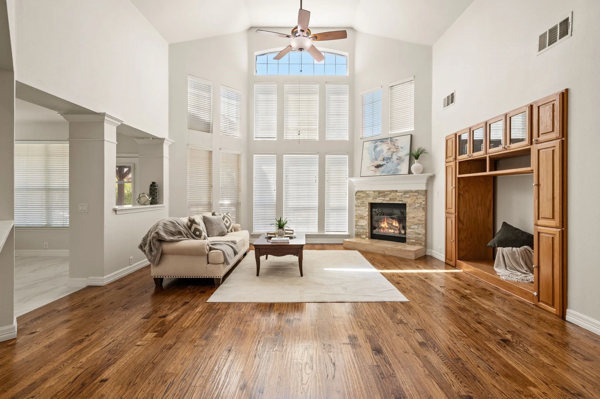 Vaulted ceiling with an integrated ceiling fan, stacked stone fireplace with a substantial hearth and mantel, two-story window wall, built-in cabinetry with glass-front uppers, and wood-finish flooring