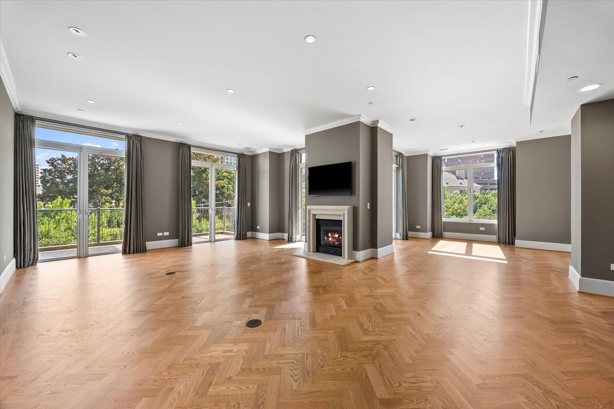 Spacious living area featuring herringbone wood-finish flooring, a contemporary fireplace with a light-toned mantel, multiple floor-to-ceiling glass doors, and recessed ceiling lighting