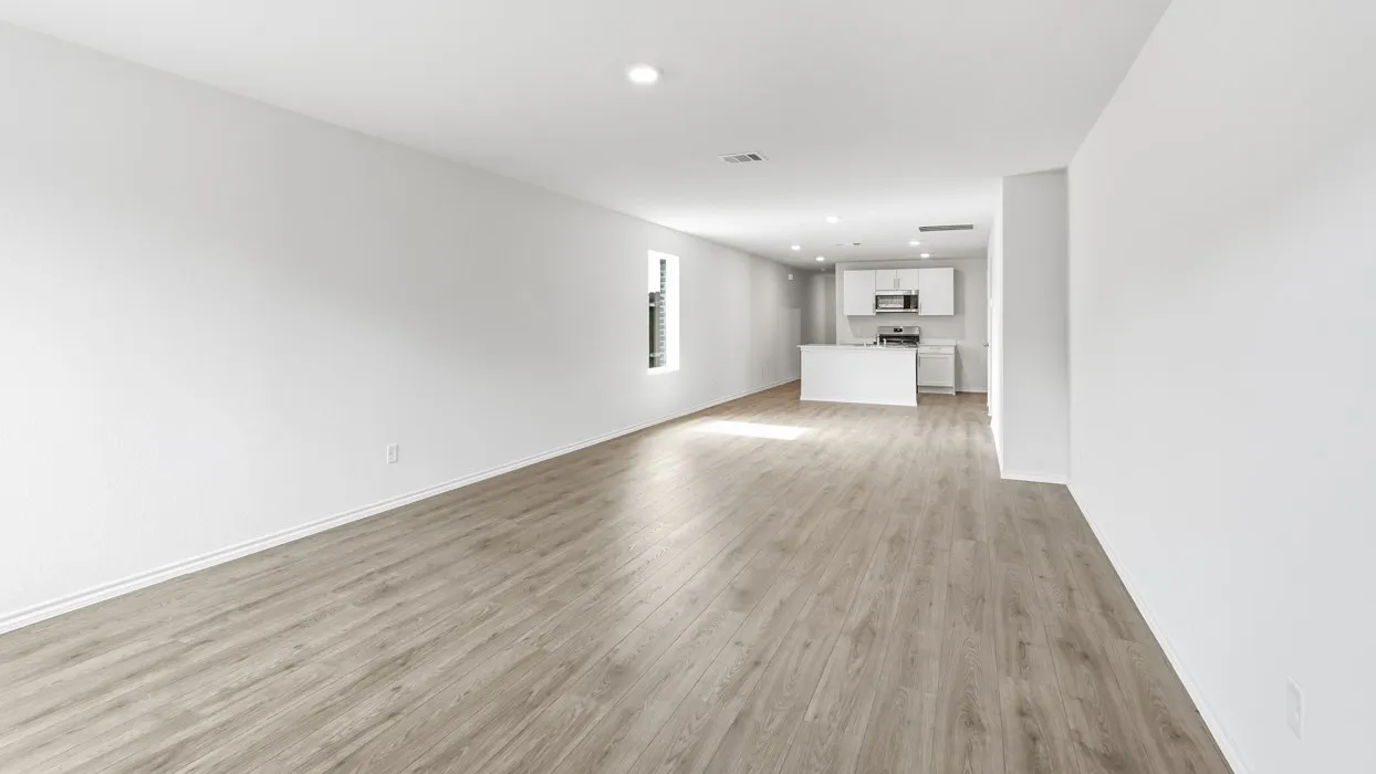 Open-concept living space featuring gleaming wood-finish flooring throughout, clean white walls, recessed ceiling lighting, and a modern kitchen with white cabinetry and stainless steel appliances visible in the background