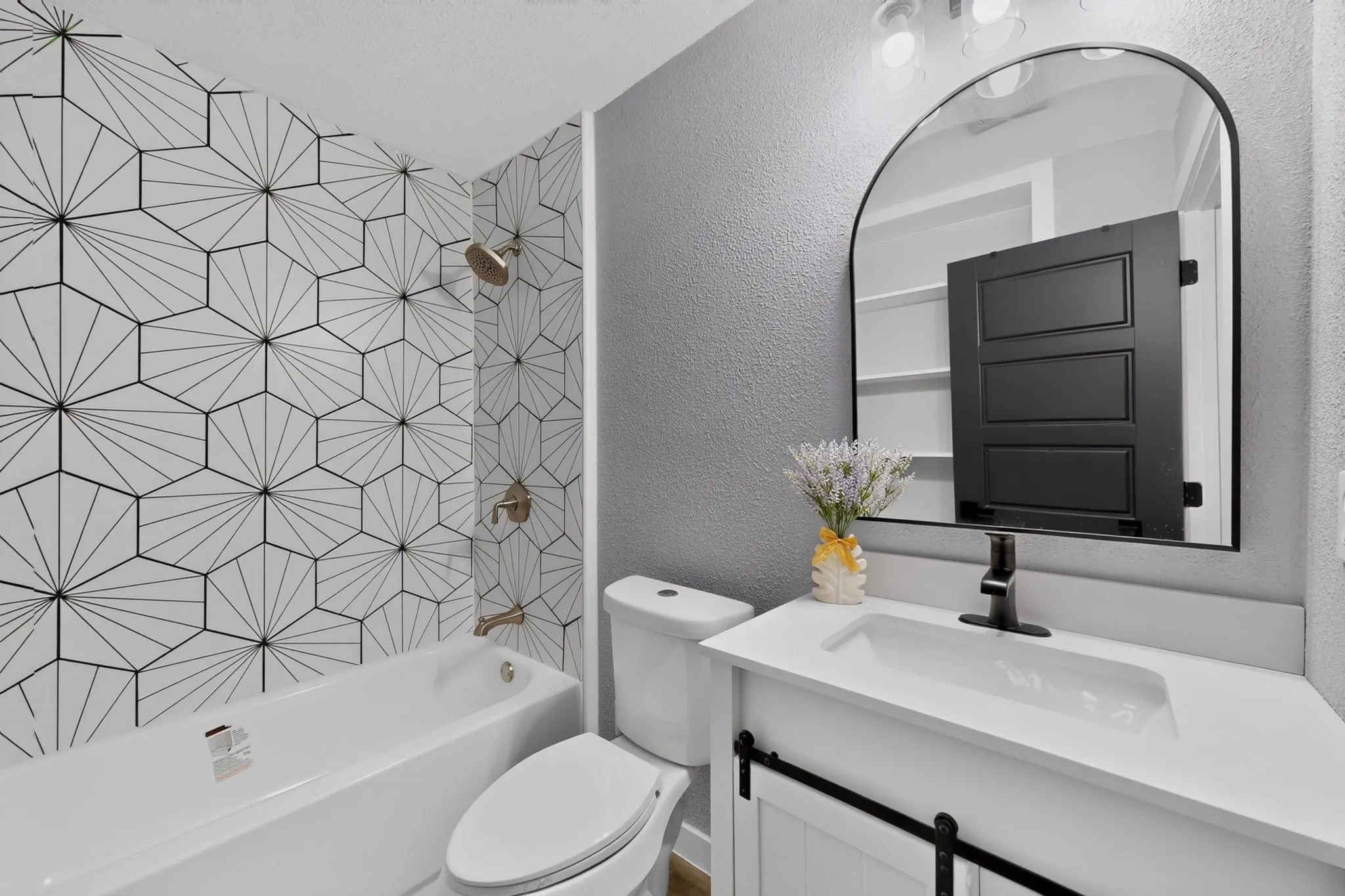 Bathtub with hexagonal patterned tile surround, white vanity with integrated sink, black barn door style vanity cabinet, arched mirror, and matte black faucet