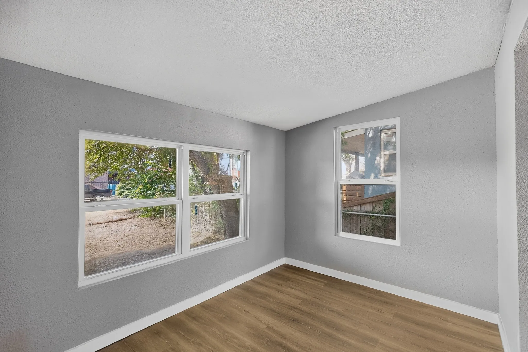 Bright corner room with dual windows framing natural light, warm wood-finish flooring, and fresh neutral wall finishes