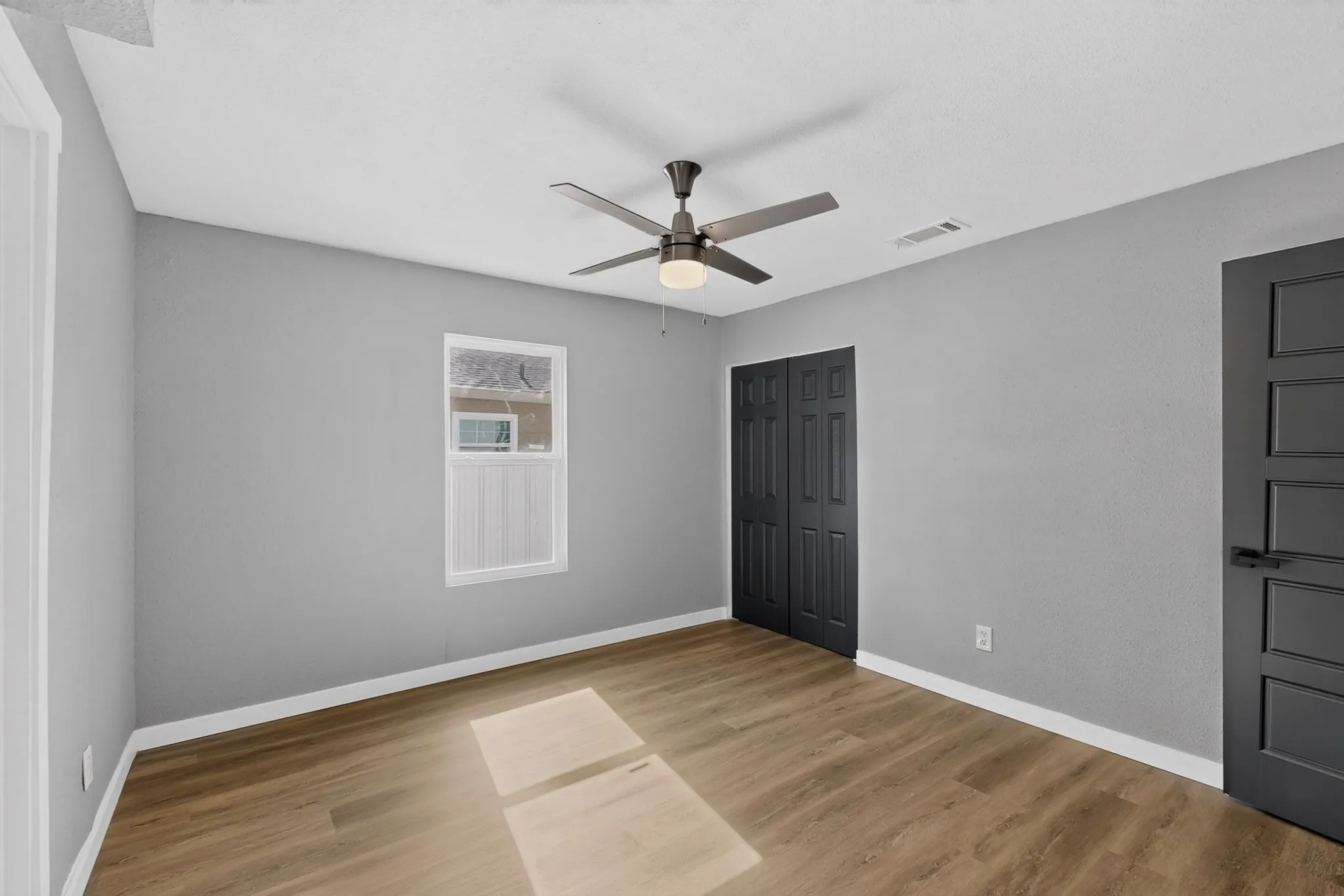 Wood-finish flooring, a ceiling fan with integrated lighting, window with white trim, and a dark-paneled closet door
