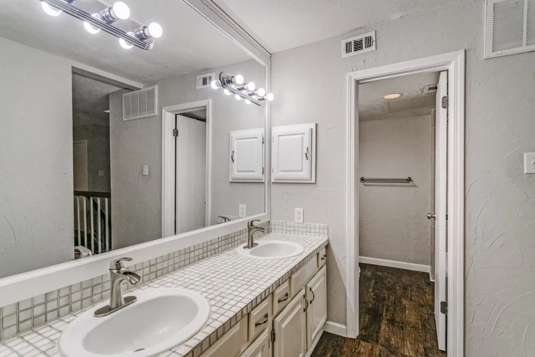 Dual undermount sinks with brushed nickel faucets, mosaic tile countertop, wood-finish flooring, and vanity lighting
