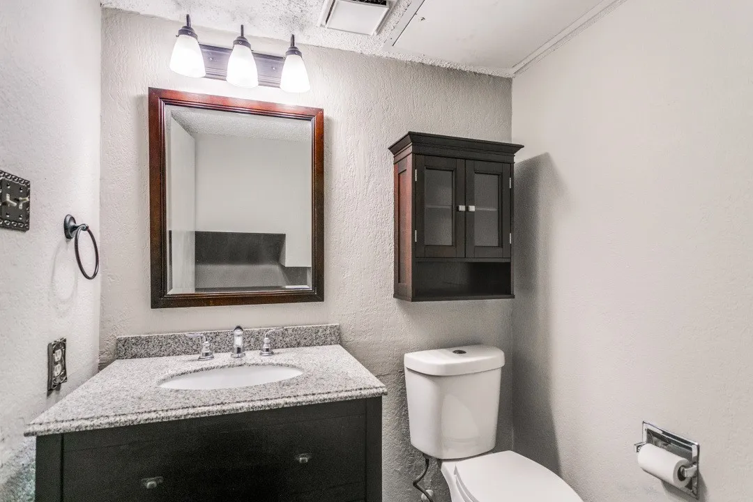 Vanity with a speckled countertop, a rectangular mirror with a wood-finish frame, a three-light vanity fixture, a dark wood-finish wall-mounted cabinet, and a white toilet