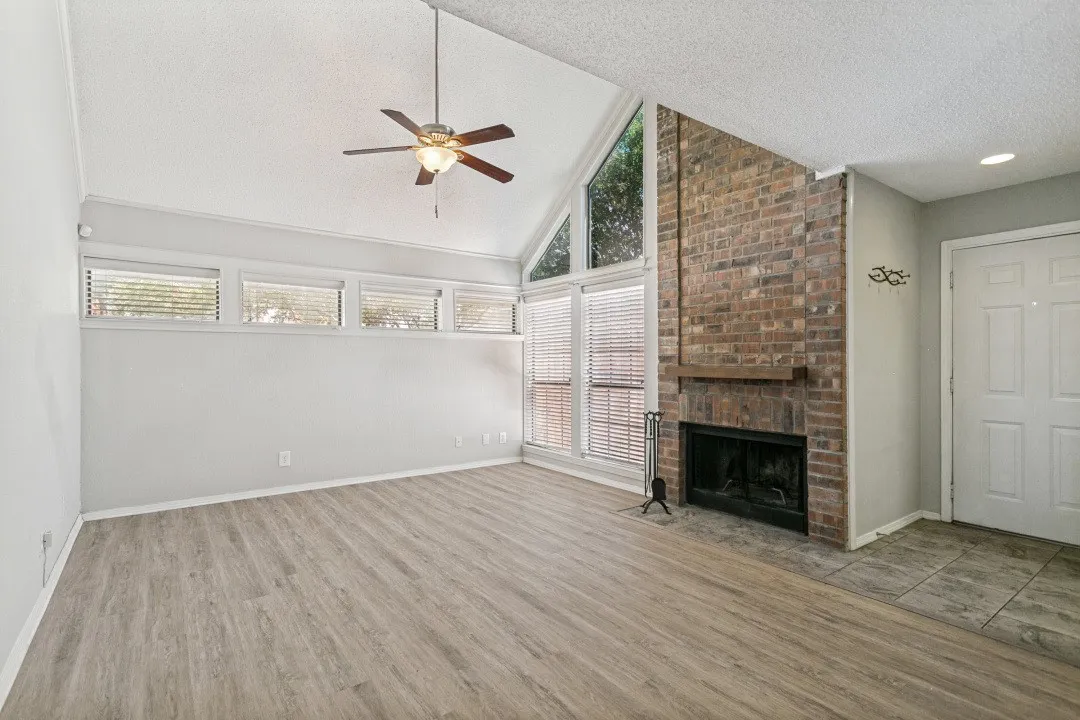 Vaulted ceiling with a ceiling fan, brick fireplace with a wood mantle, wood-finish flooring, and multiple large windows