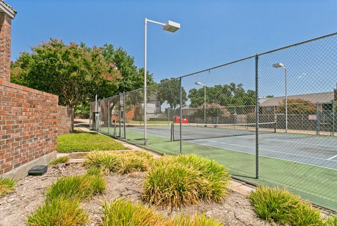 Tennis court with chain-link fencing, featuring overhead lighting fixtures and a brick wall perimeter