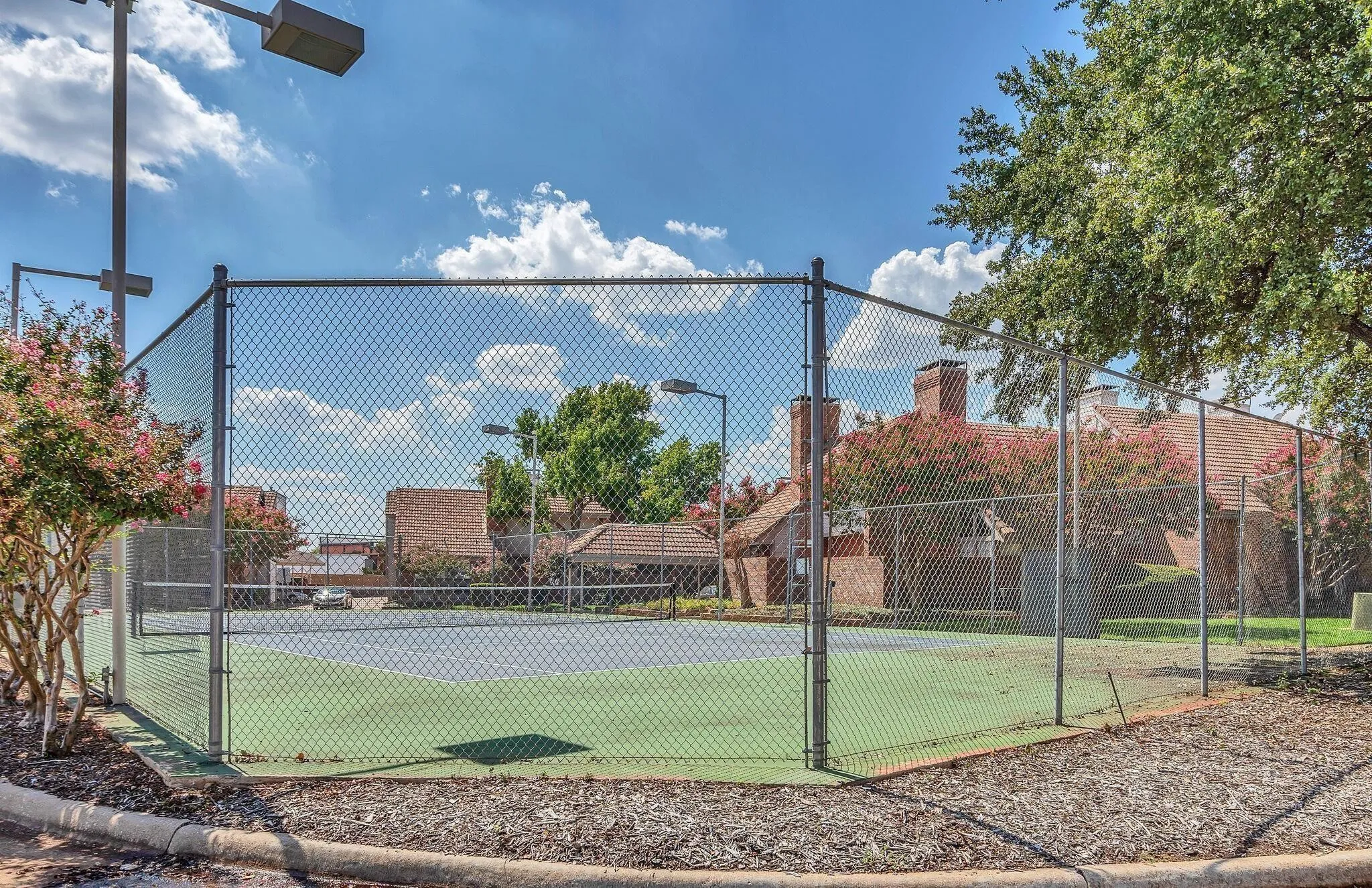 Outdoor tennis court with chain-link fencing, featuring light poles and a concrete curb