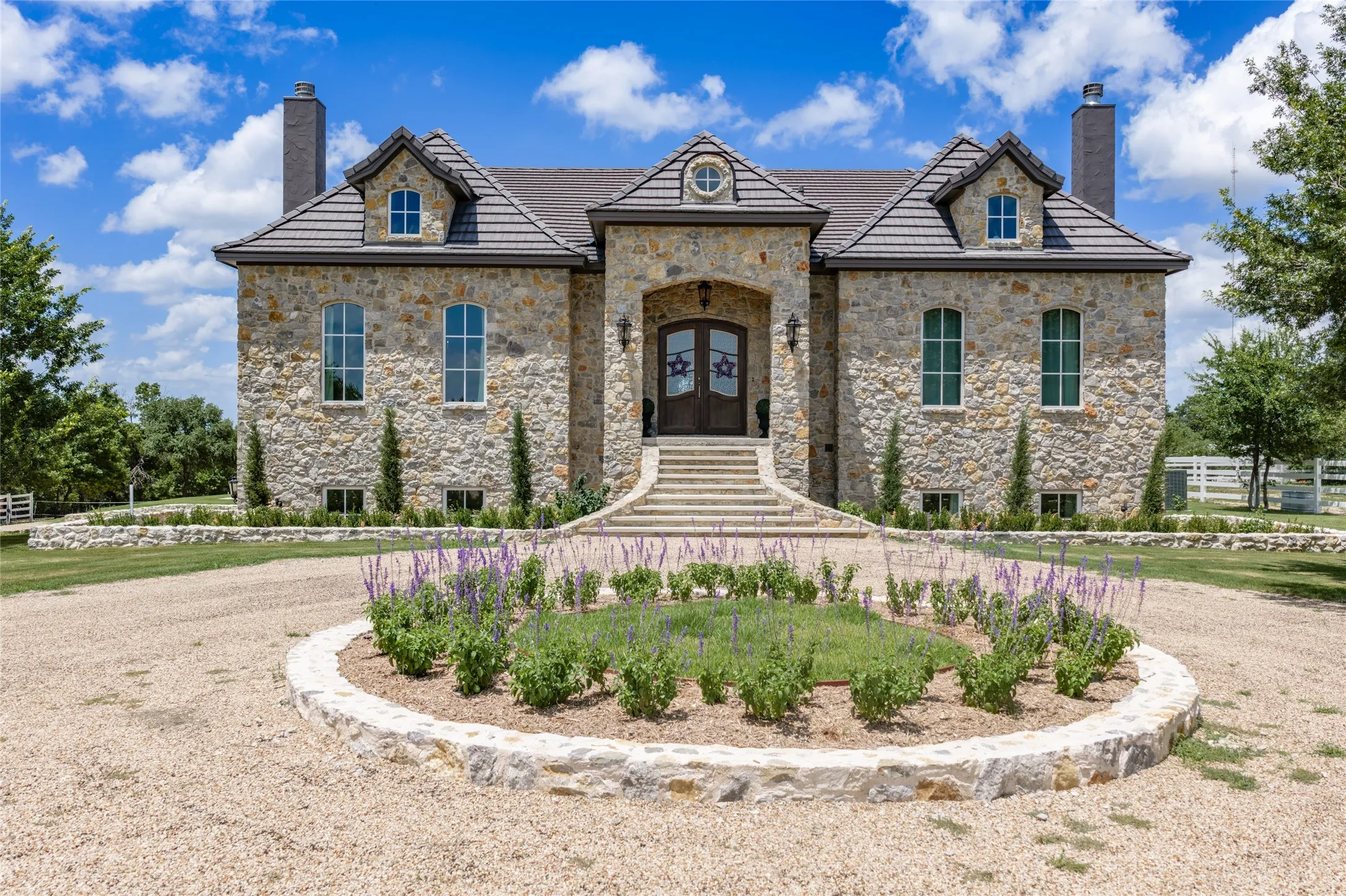 Stone facade with a multi-level roofline, arched entry, dormer windows, and a circular gravel driveway featuring a landscaped island