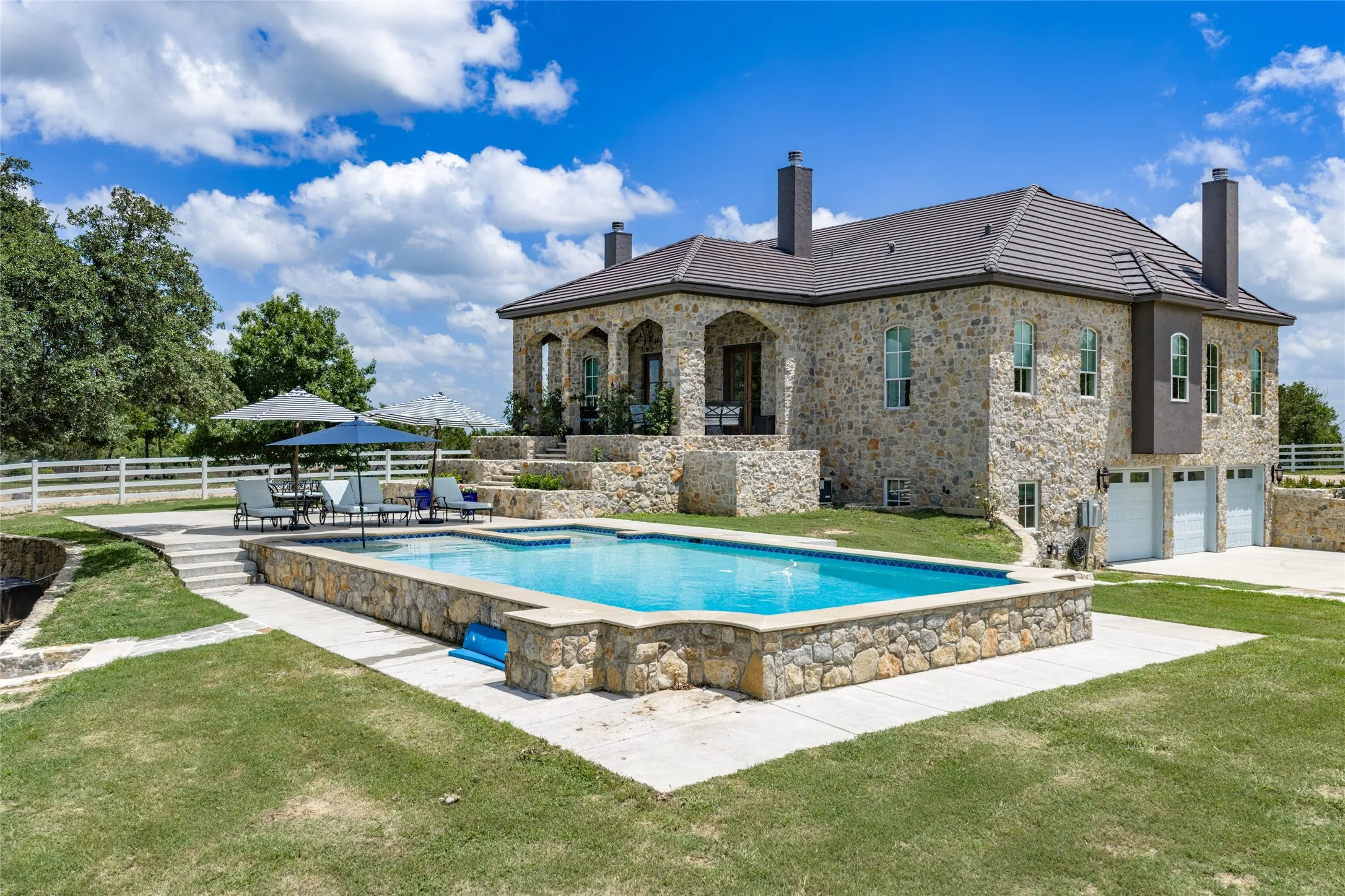 Stone exterior residence featuring a tiled roof, multi-car garage, tiered outdoor living spaces, a rectangular swimming pool, and white fencing