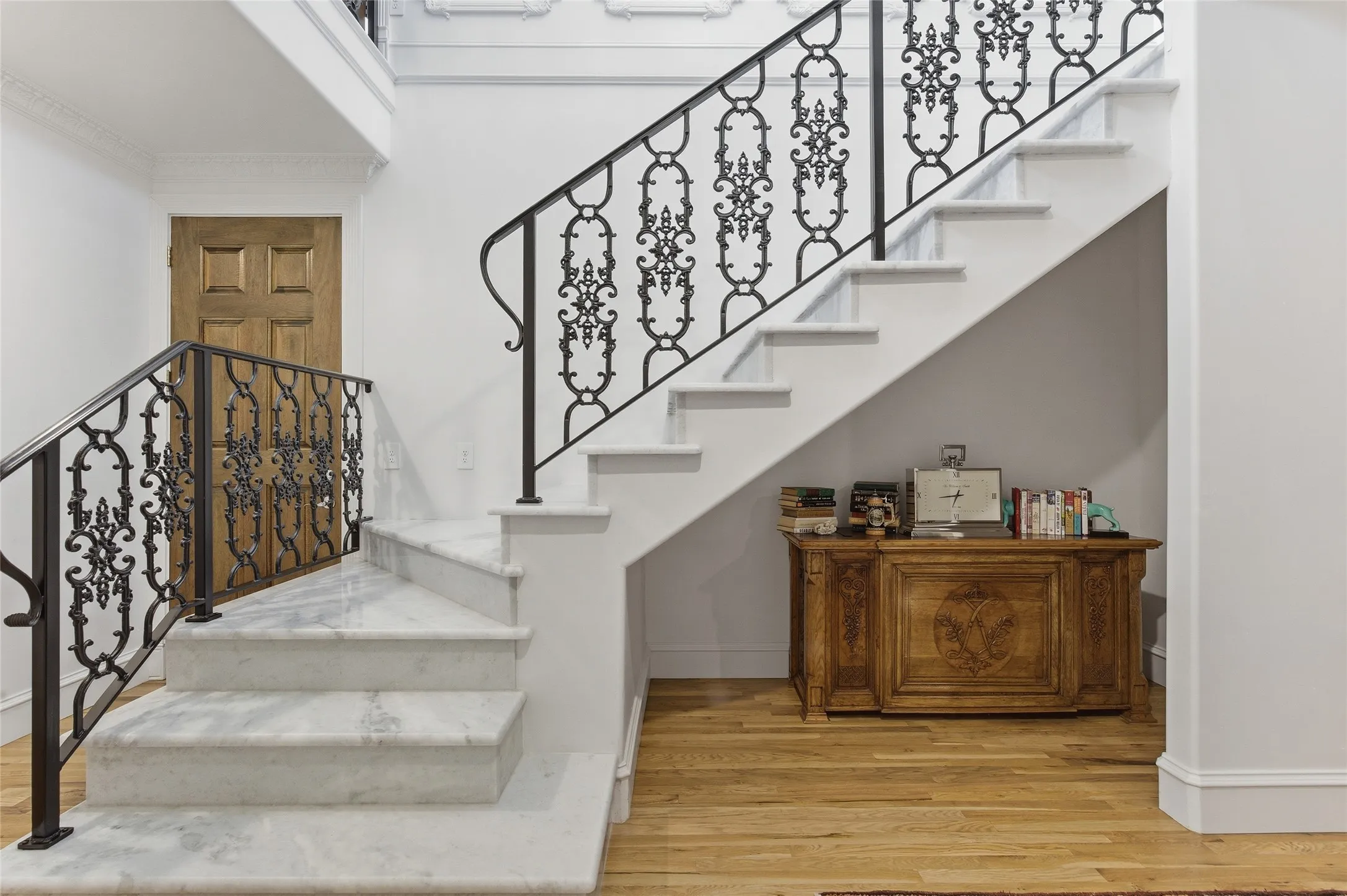 White marble staircase with ornate black wrought iron railings, light wood-finish flooring, and white crown molding