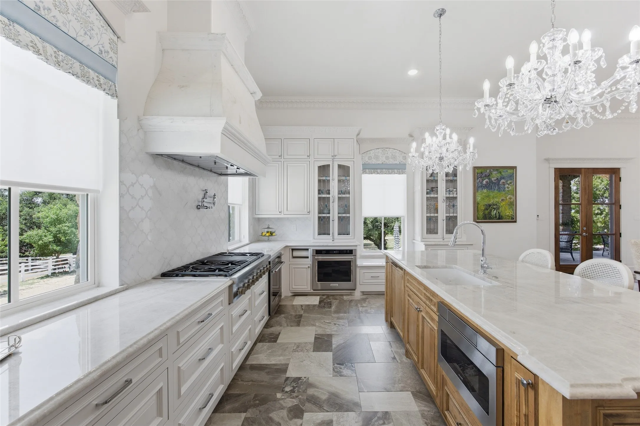 Gourmet kitchen featuring a built-in range, custom cabinetry, large central island, overhead crystal chandeliers, and patterned tile flooring