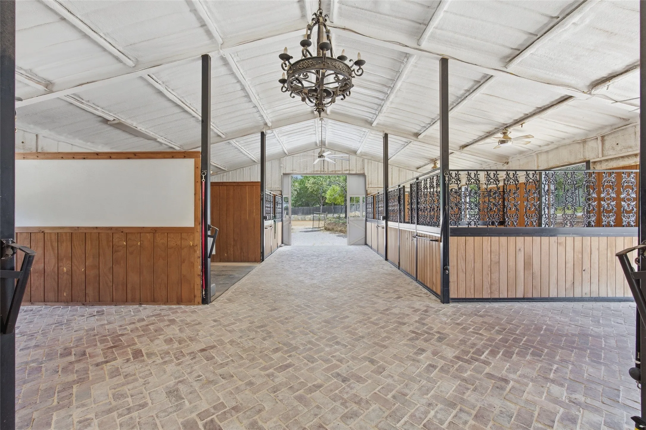 Expansive interior featuring a herringbone brick floor, exposed beam ceiling, wrought iron chandelier, wood-paneled walls, and built-in stalls with decorative metalwork