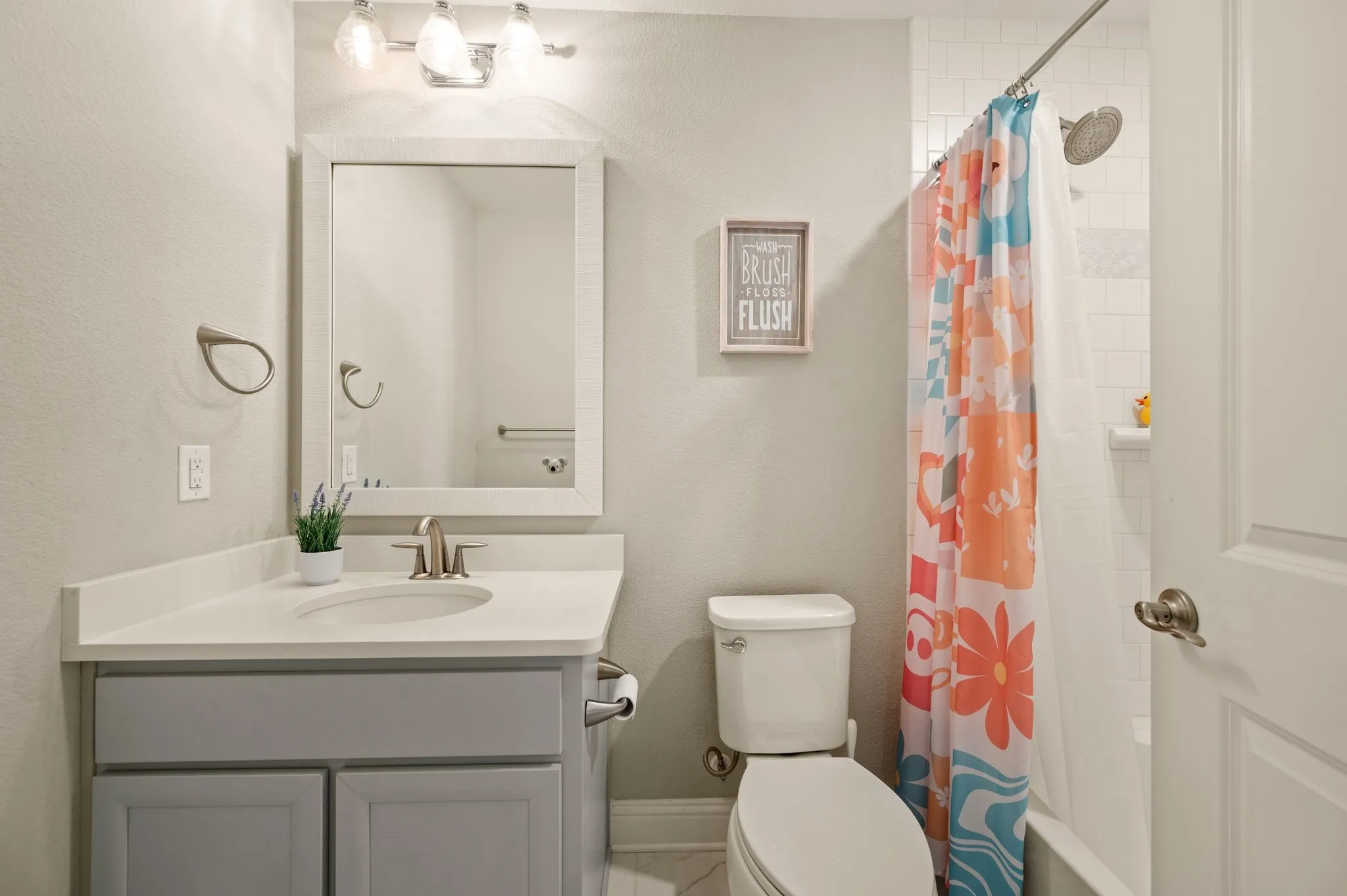 Bright bathroom featuring a gray vanity cabinet with ample storage, white quartz countertop, large framed mirror with integrated lighting, and brushed nickel fixtures