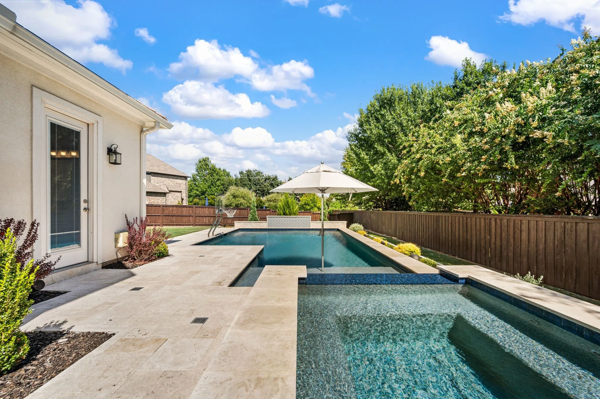 Resort-style backyard oasis featuring a sparkling saltwater pool with integrated spa, travertine patio decking, mature landscaping with privacy fencing, pergola shade structure, and direct access from the home's exterior