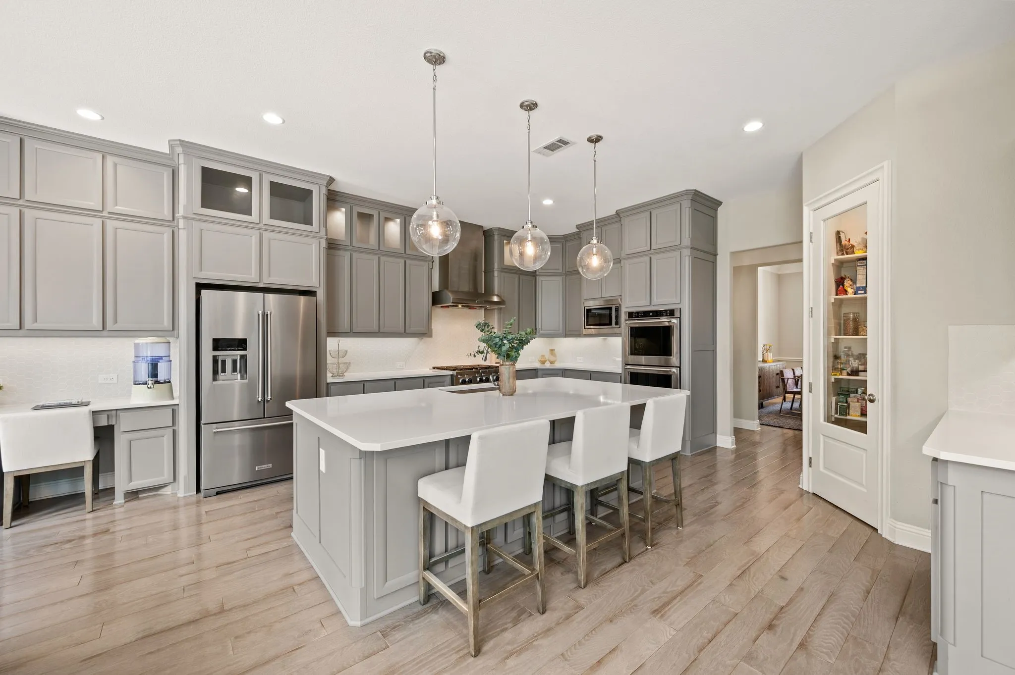 Chef's kitchen featuring custom cabinetry in soft gray and white finishes, stainless steel appliances including a built-in refrigerator and wall-mounted oven, white quartz countertops, and a large center island with seating