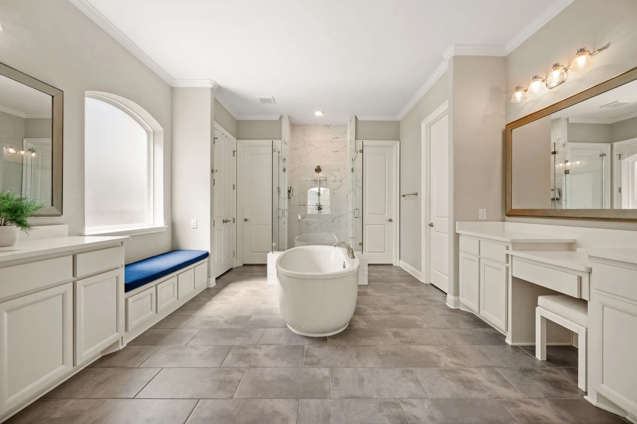 Freestanding soaking tub, dual vanities with white cabinetry, glass-enclosed shower, large arched window, and tiled flooring