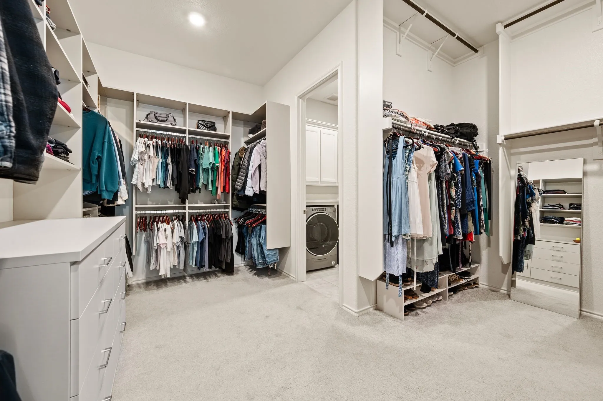 Spacious walk-in closet with custom shelving and hanging systems, light-finish cabinetry with multiple drawers, neutral carpeting, and integrated washer/dryer appliance for added convenience