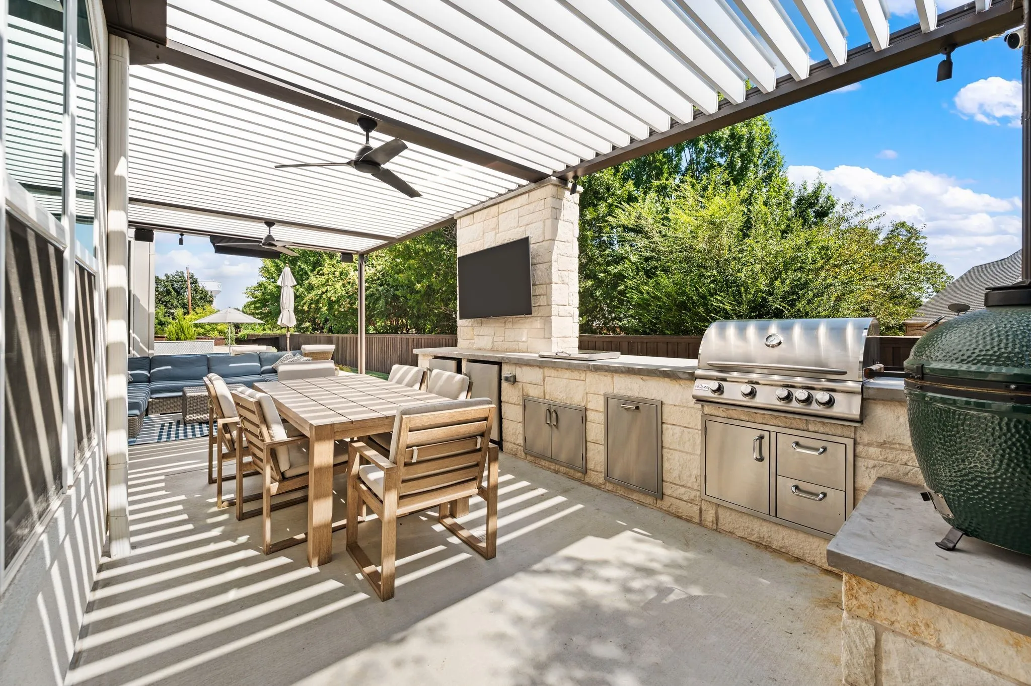Sophisticated outdoor entertaining space featuring a motorized louvered pergola with integrated ceiling fan, custom stone countertops and cabinetry with stainless steel appliances including a built-in grill, dining table with seating for six, and mature landscape screening
