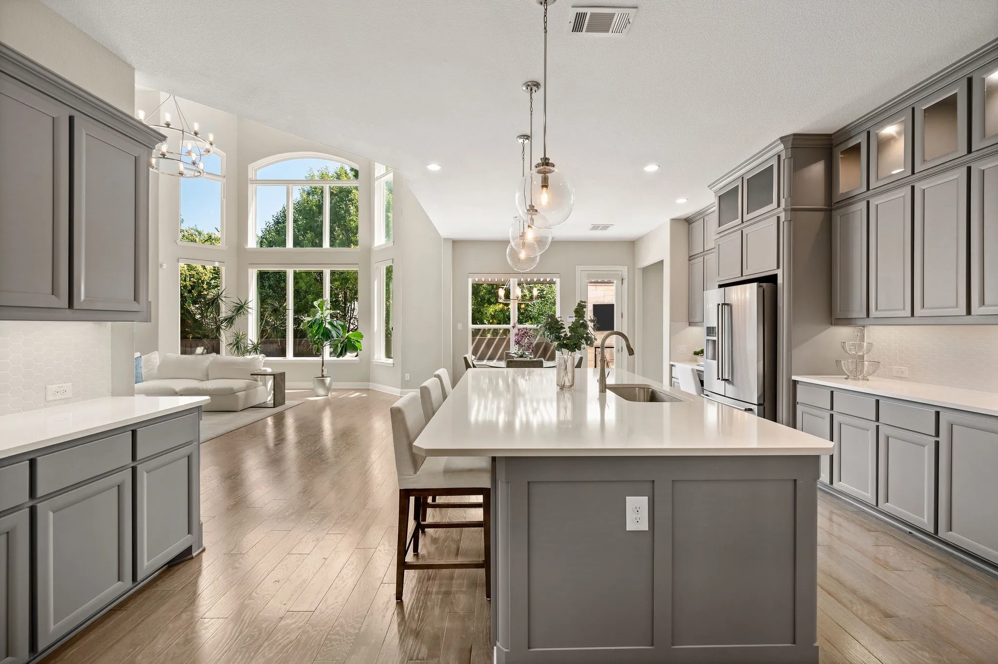Gourmet kitchen featuring gray cabinetry with shaker-style doors, white quartz countertops, a large center island with seating, stainless steel appliances, pendant lighting, and abundant natural light from oversized windows overlooking the grounds